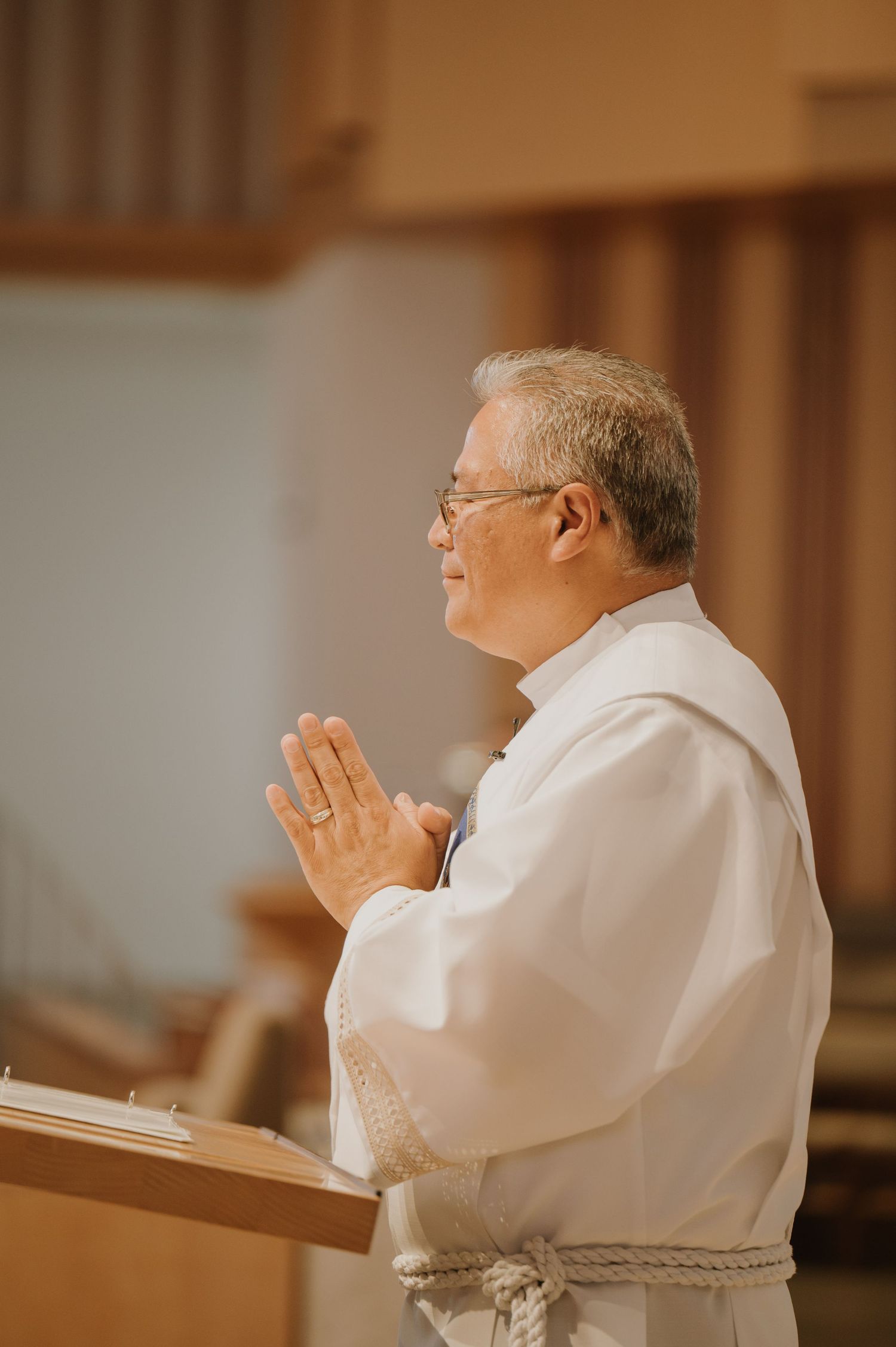 Smiling priest in white vestments prays with hands together at church wedding ceremony altar.
