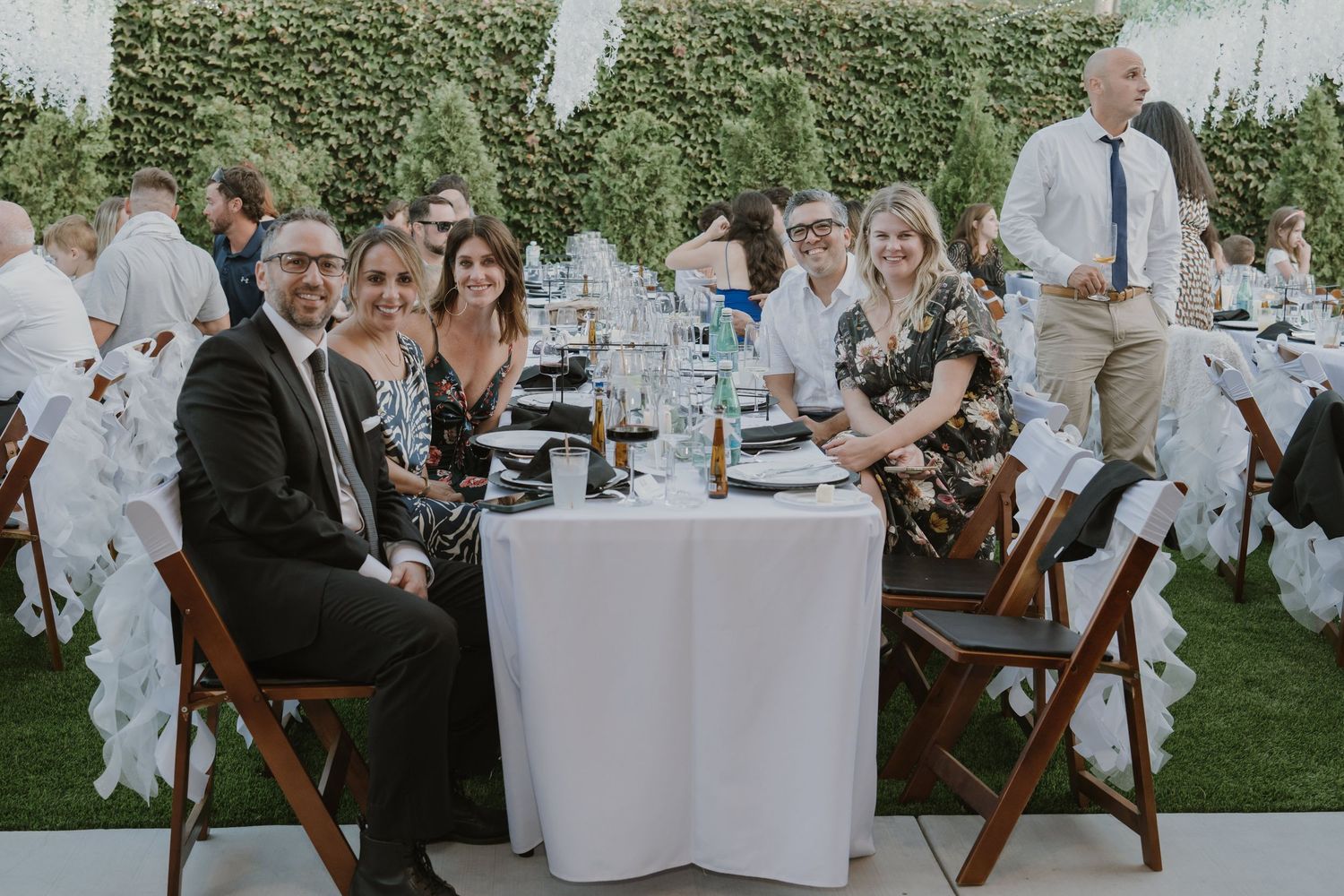 Wedding guests smile and pose at decorated reception table during outdoor garden wedding celebration.