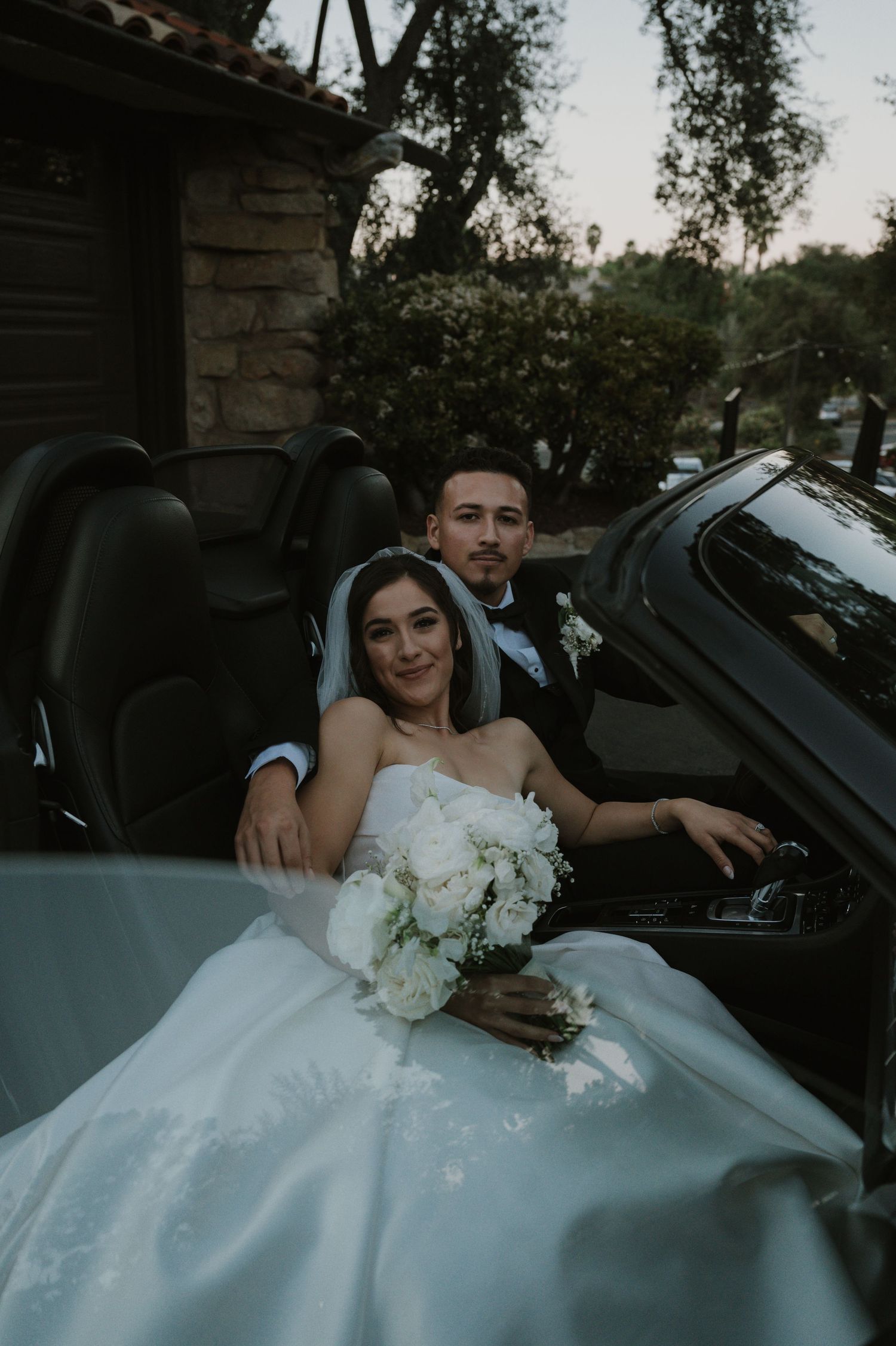 A couple poses together in a convertible car with a white wedding bouquet.