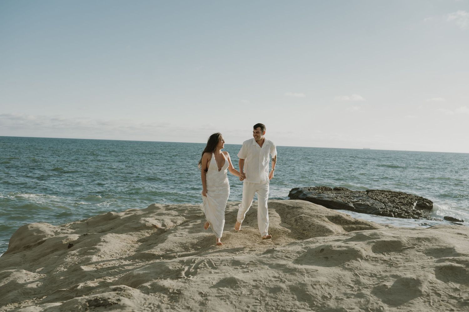 Two people in white clothing walking along dramatic ocean cliffs at sunset.