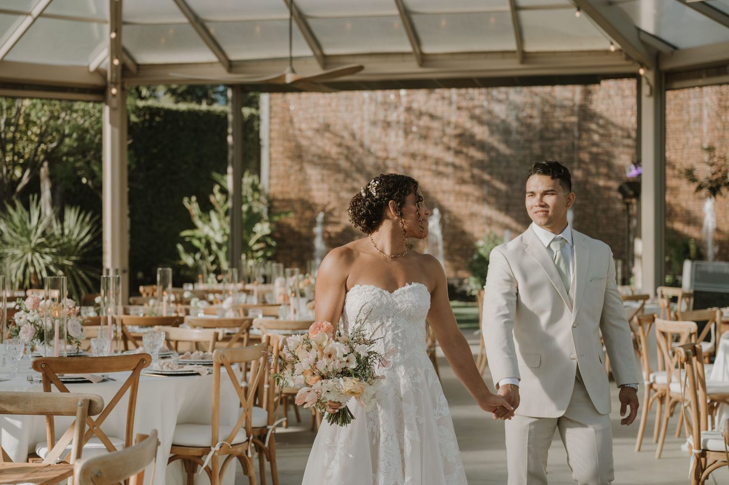 Bride and groom walk hand in hand in an elegant glass-roofed wedding venue with wooden chairs and floral centerpieces.
