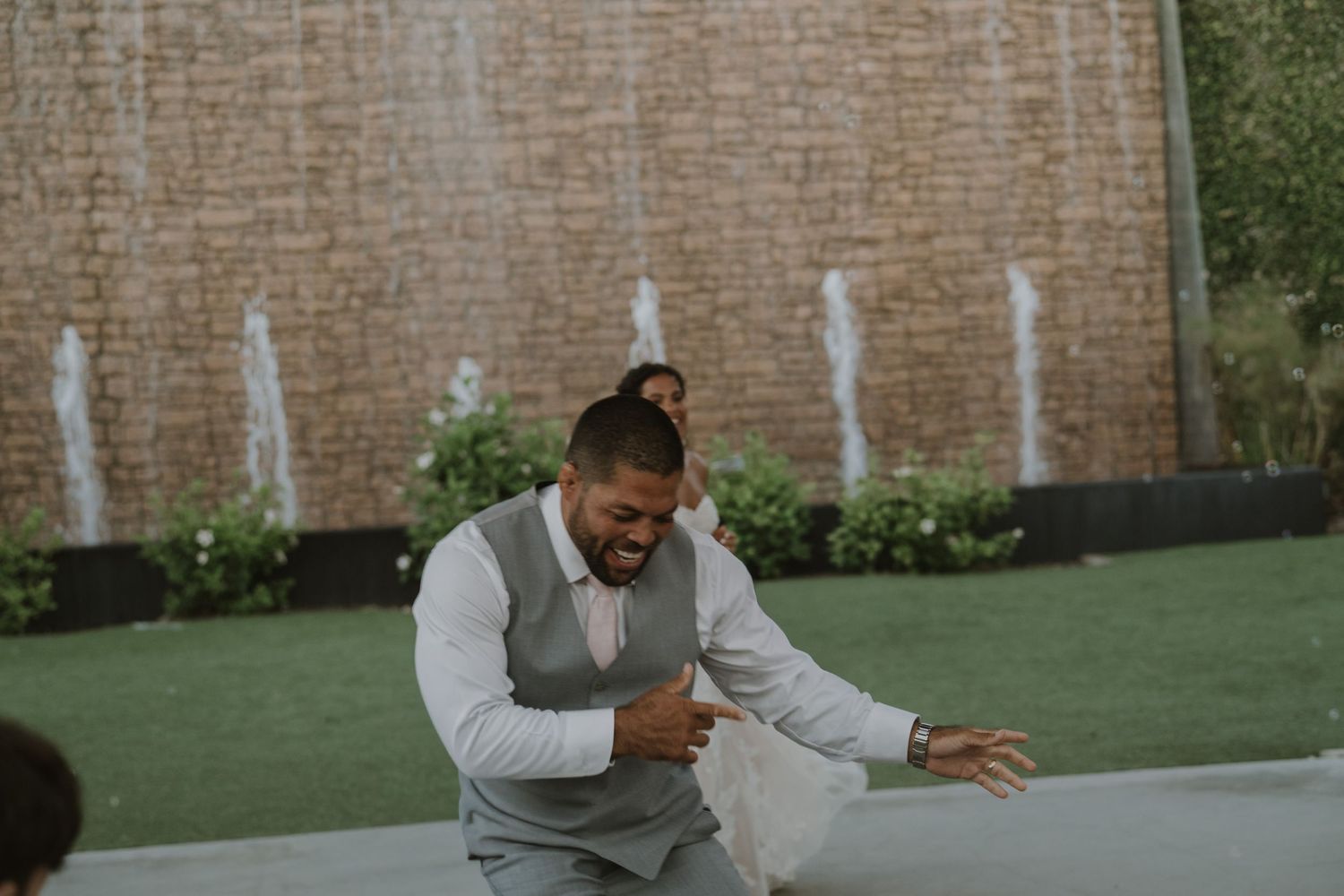 Groom dancing joyfully outdoors near a decorative water fountain wall.
