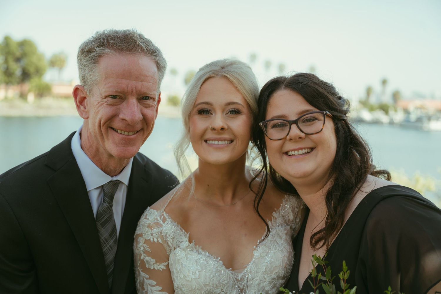 Bride in lace gown smiles with two guests at outdoor waterfront wedding celebration.