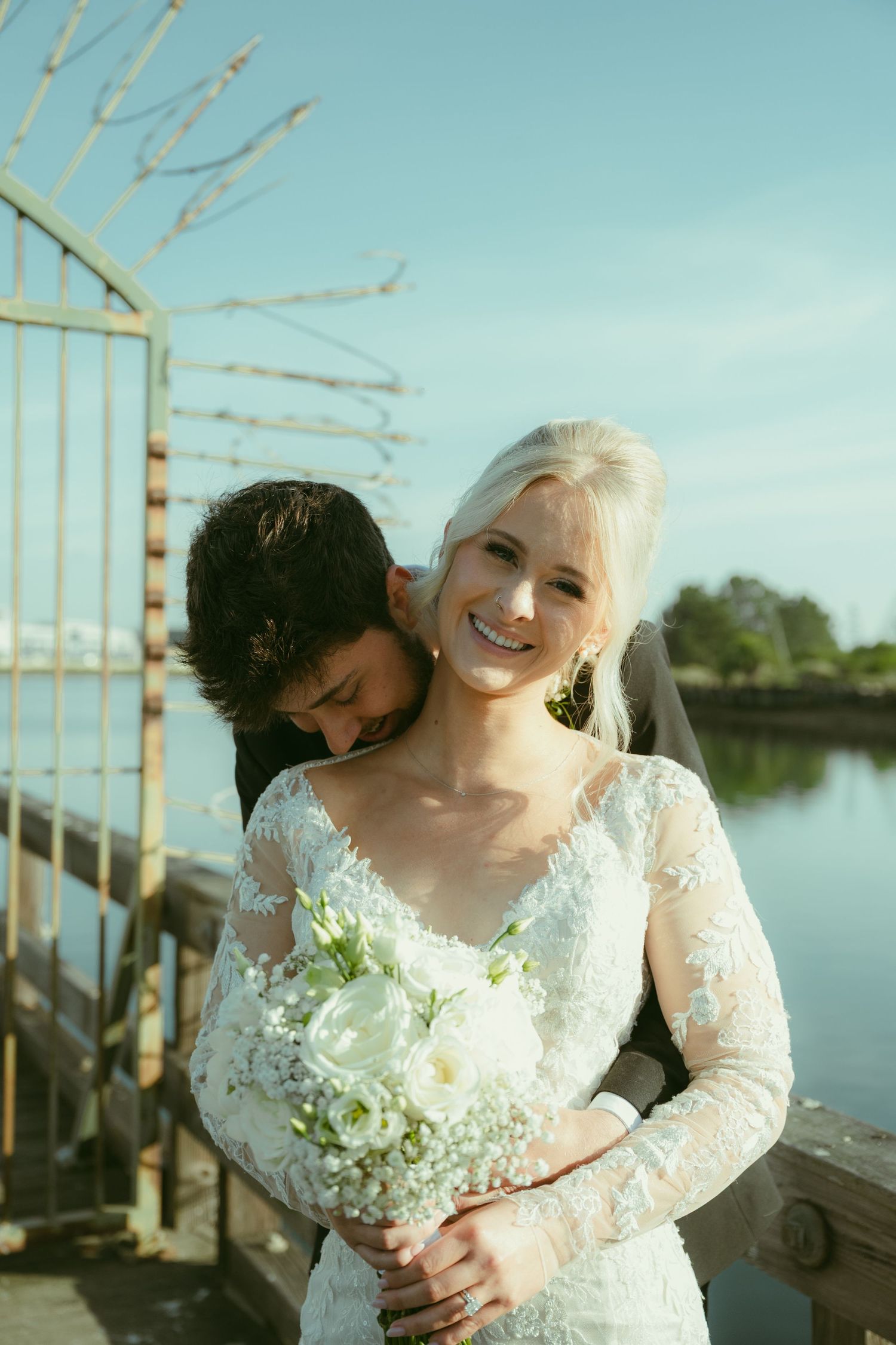 Smiling bride holds white bouquet as groom embraces her by a sunny waterfront.
