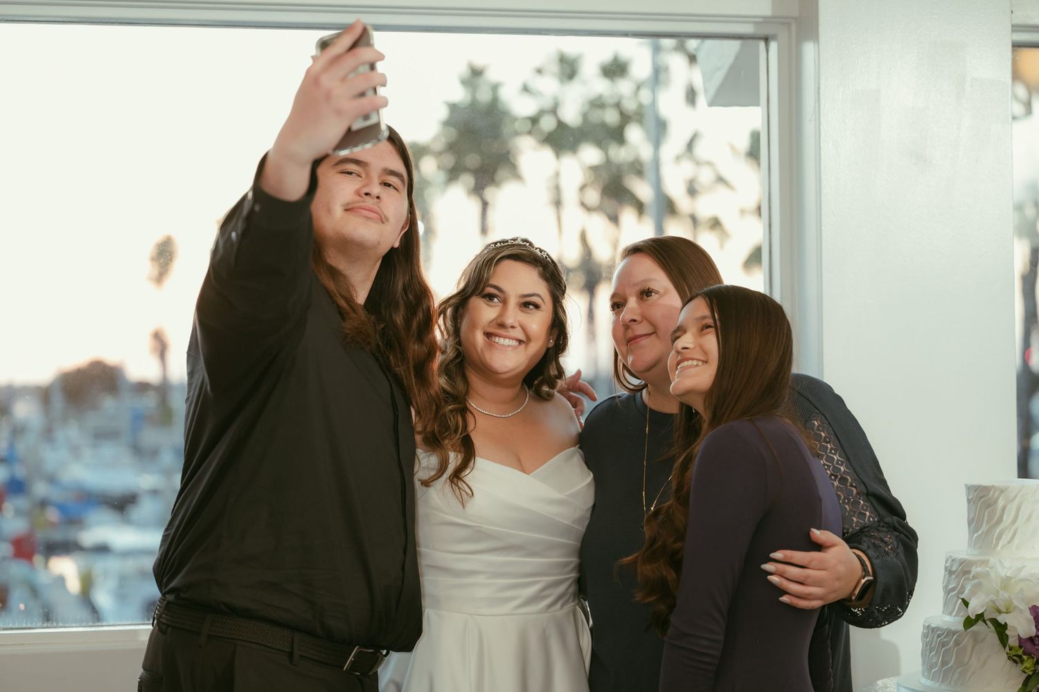 Bride takes a selfie with family members at a waterfront wedding venue with palm trees visible outside.