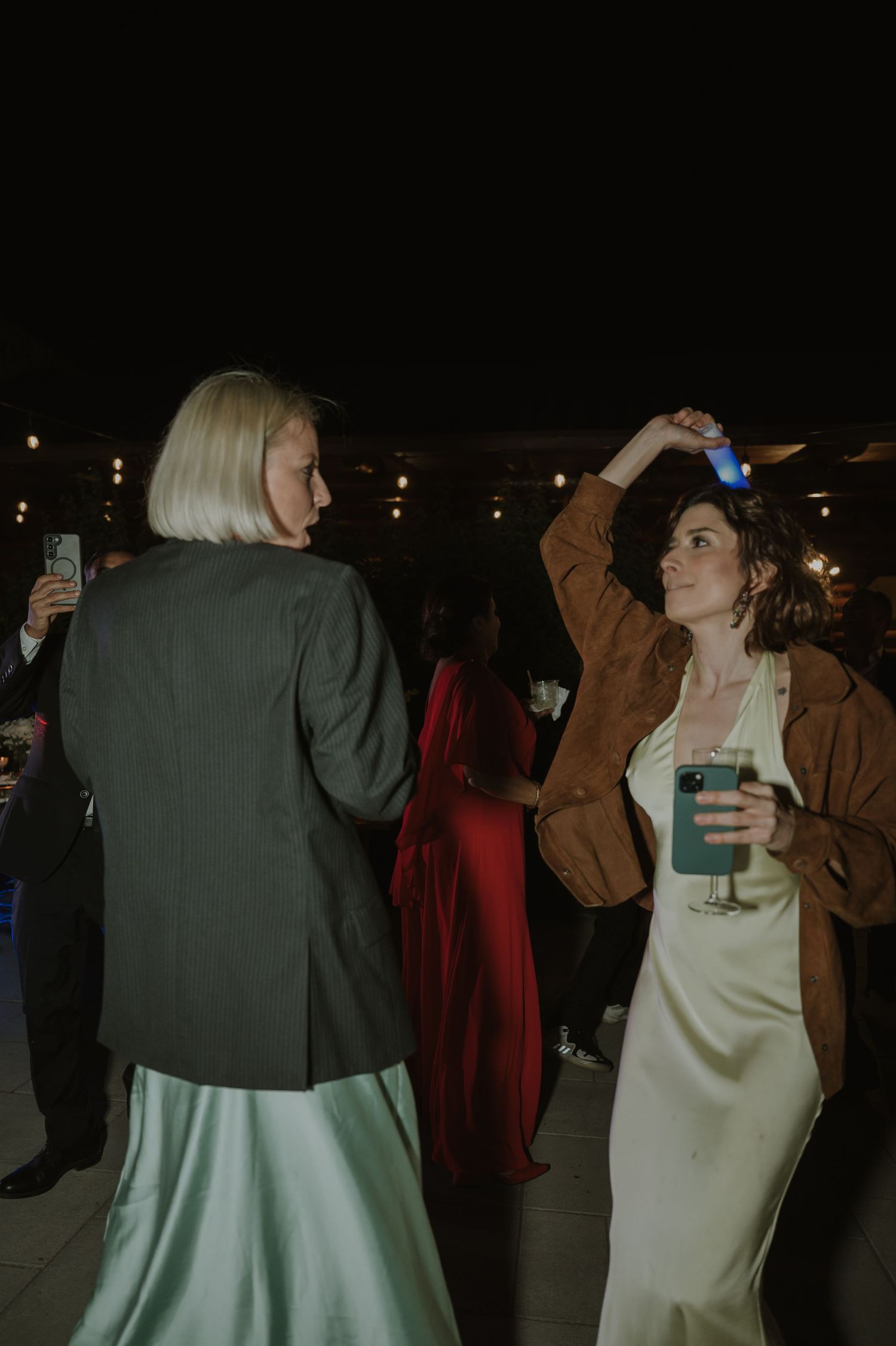 Two women dancing at a wedding reception at night, one in a green satin dress with a blazer holding a drink.