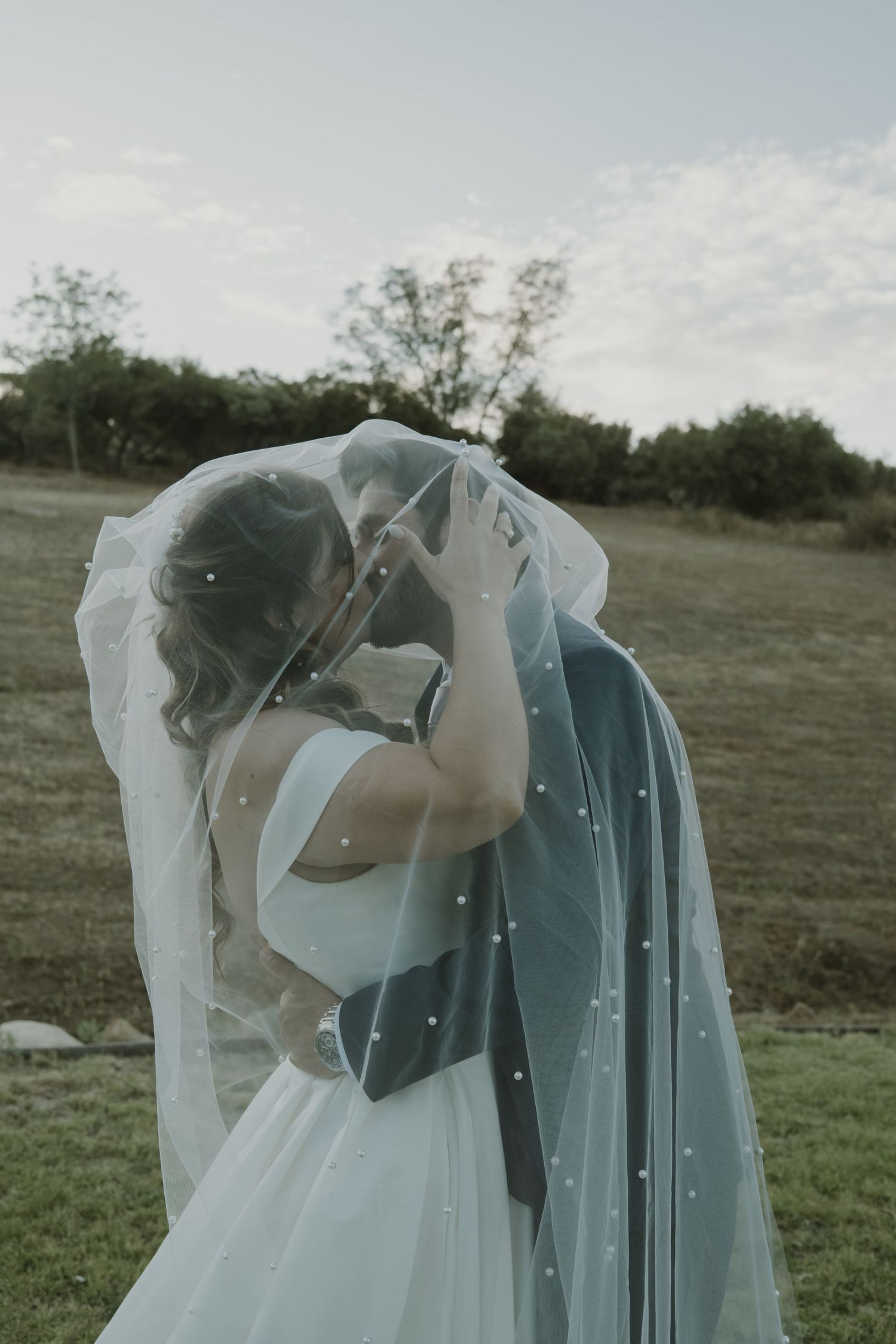 Romantic bride and groom sharing a kiss under a pearl-adorned wedding veil in an outdoor setting.