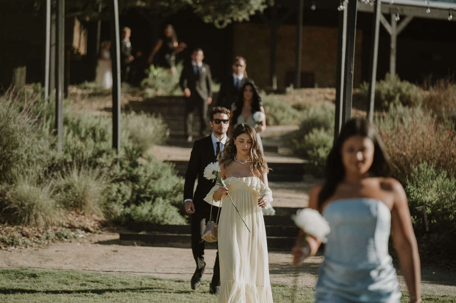 Wedding party walking along garden path with bride in white dress holding bouquet at outdoor venue.