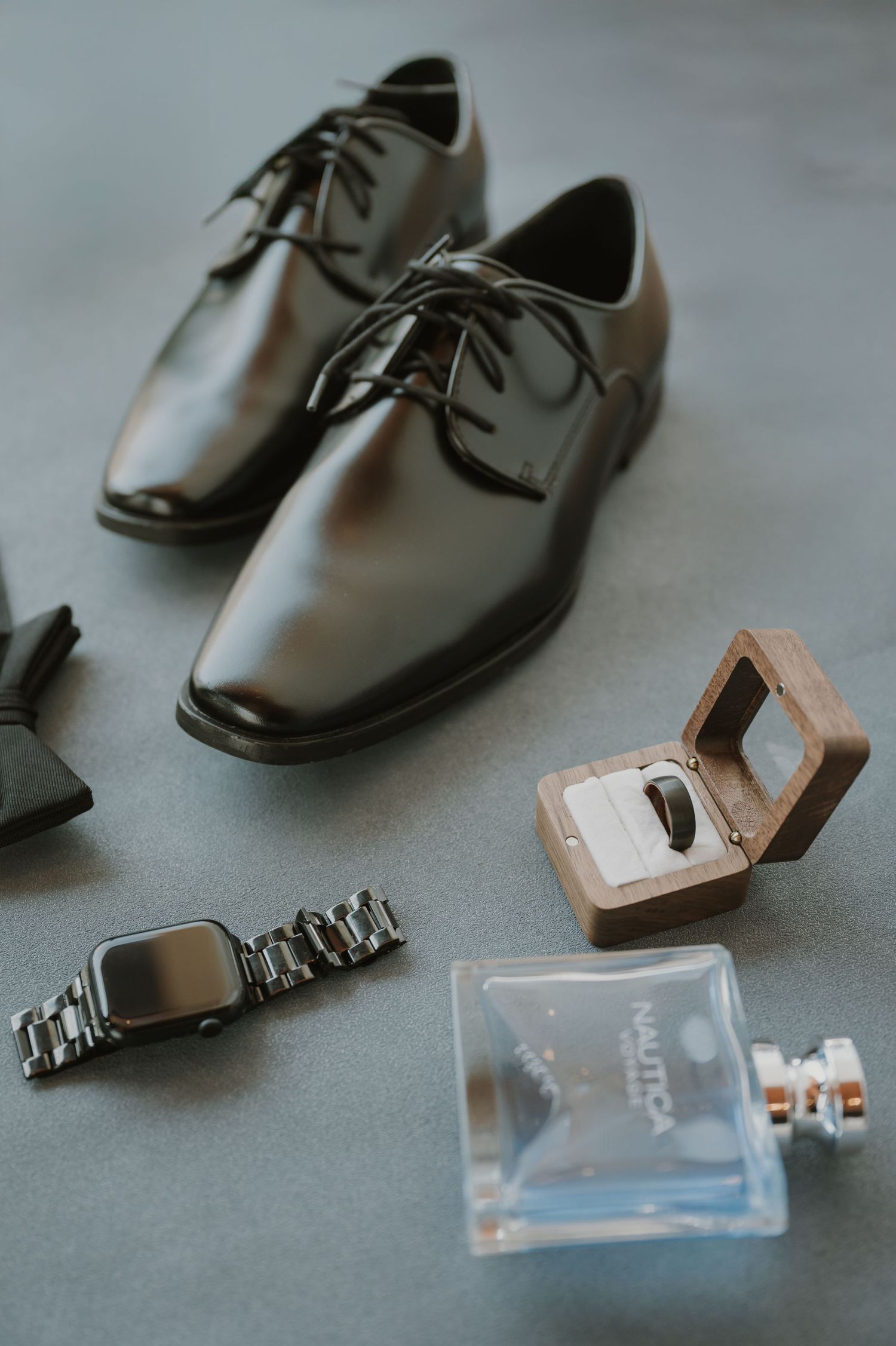 Black dress shoes, bow tie, silver watch, wooden ring box, and Nautica cologne arranged as groom wedding accessories.