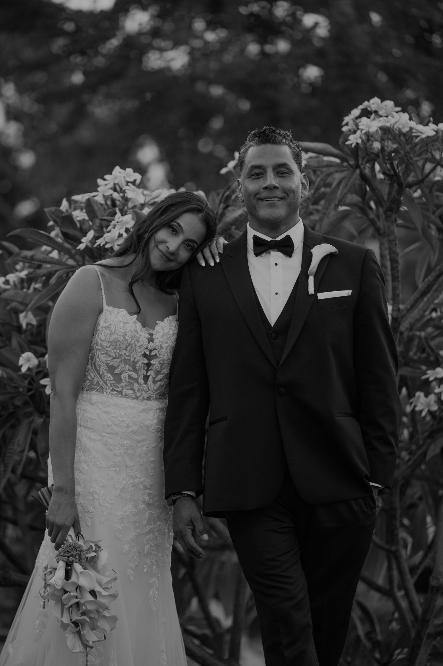 A bride and groom pose together in a black and white portrait surrounded by lush flowers.