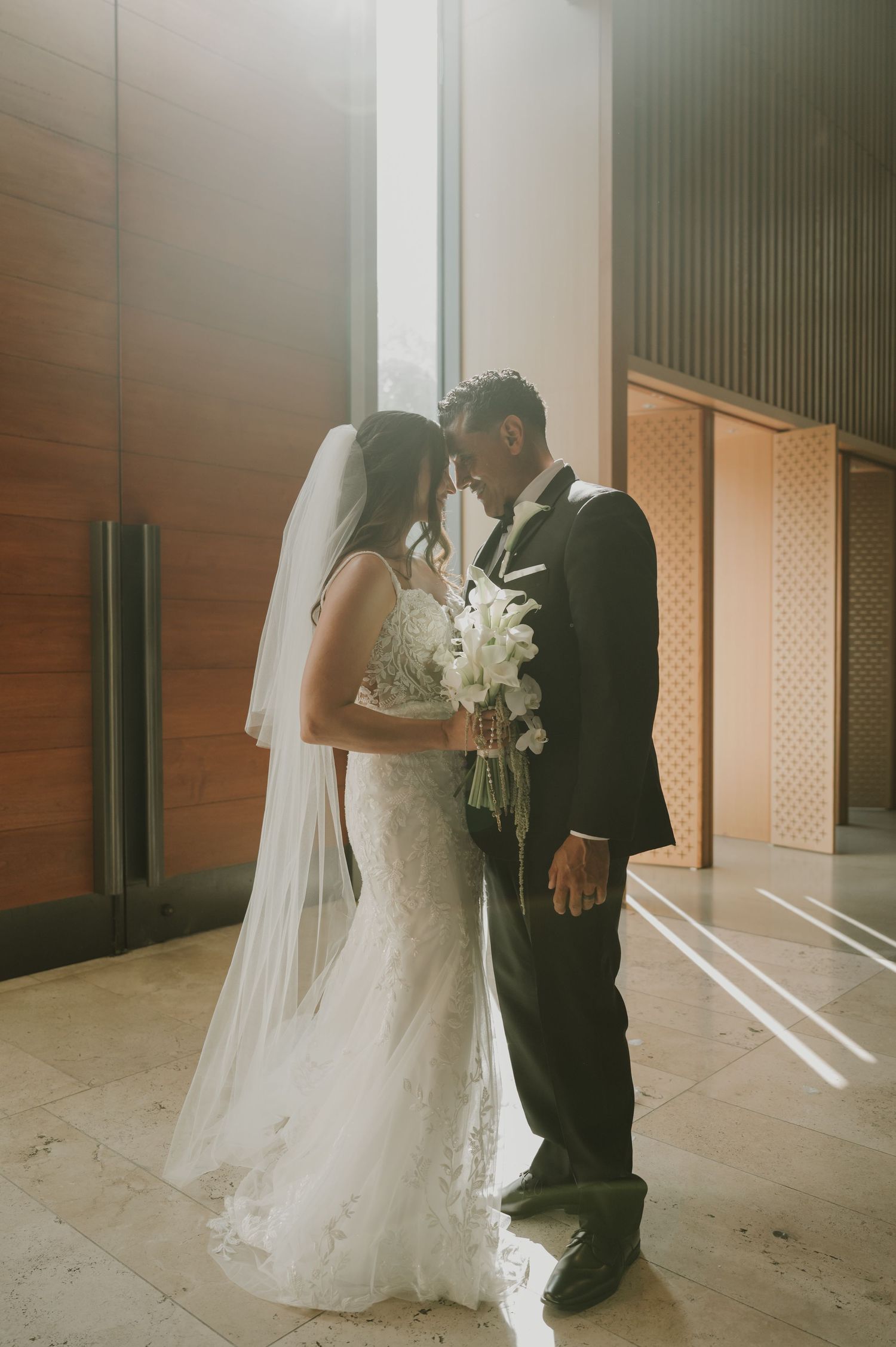 Bride in lace gown and veil holds white bouquet as couple touches foreheads in a sunlit modern venue.