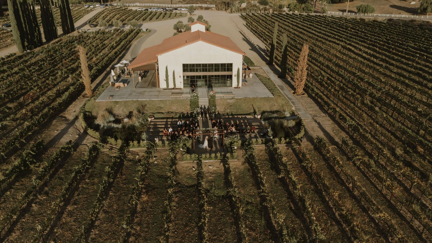 Aerial view of a white wedding venue building surrounded by vineyard rows.