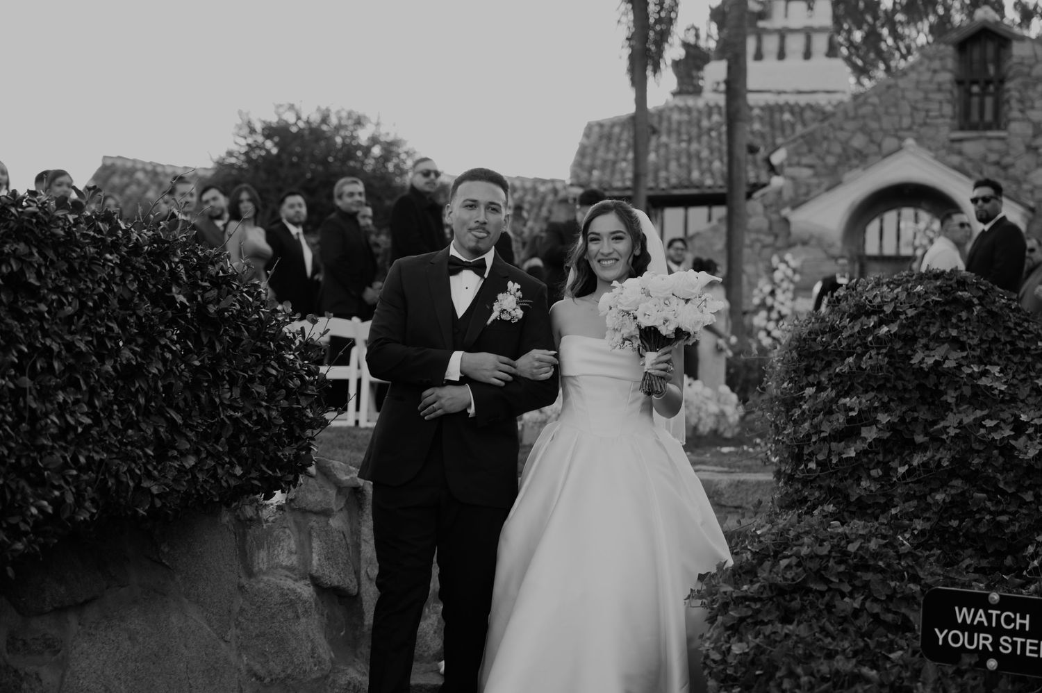 A wedding couple walking through a garden path with stone architecture in the background.