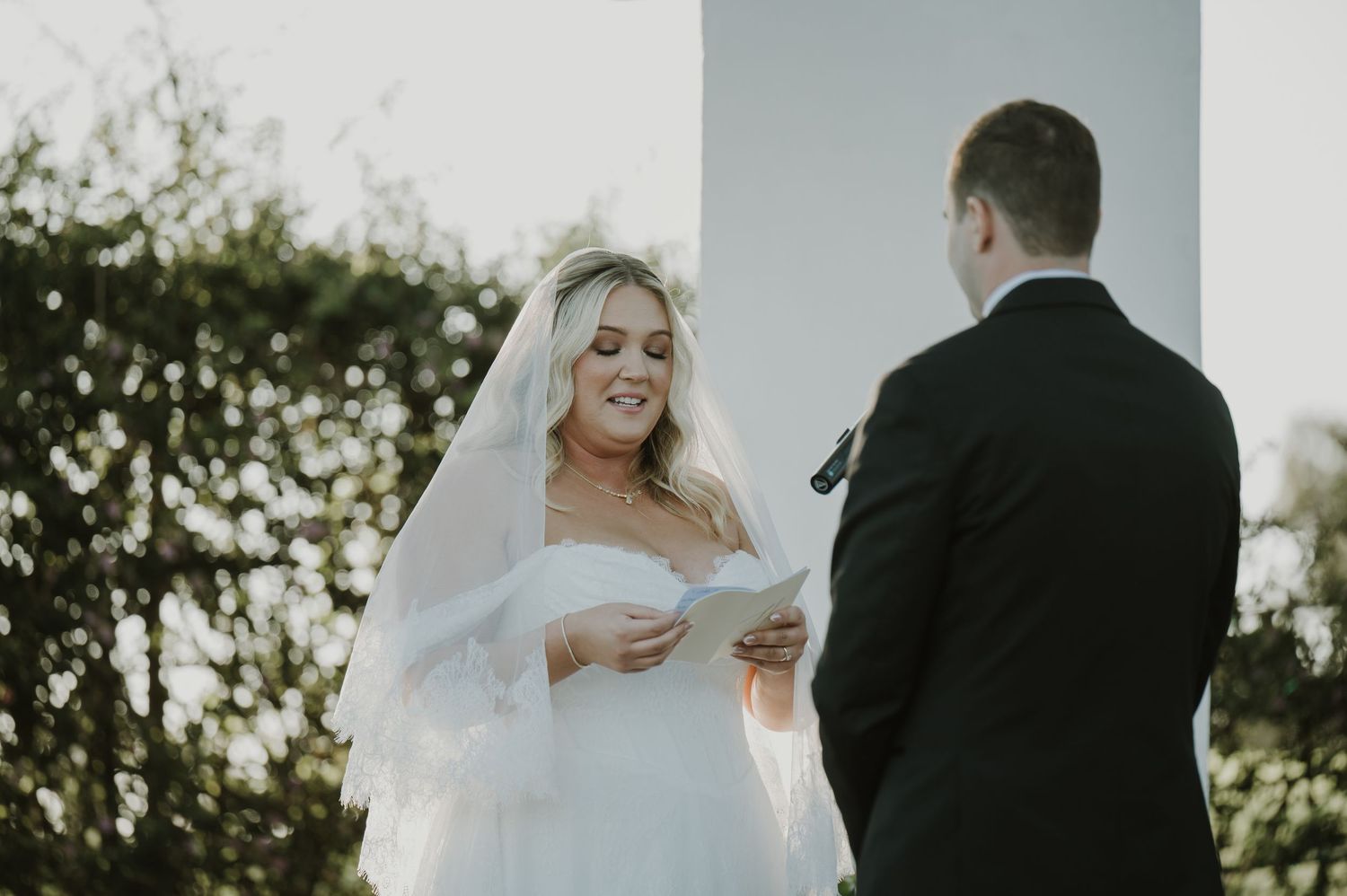 Smiling bride in white veil reads heartfelt wedding vows to groom at outdoor ceremony.