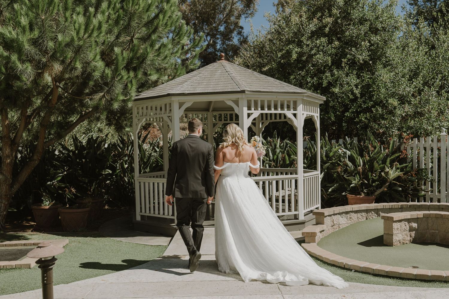 Bride and groom walk hand in hand toward a white garden gazebo surrounded by lush greenery.