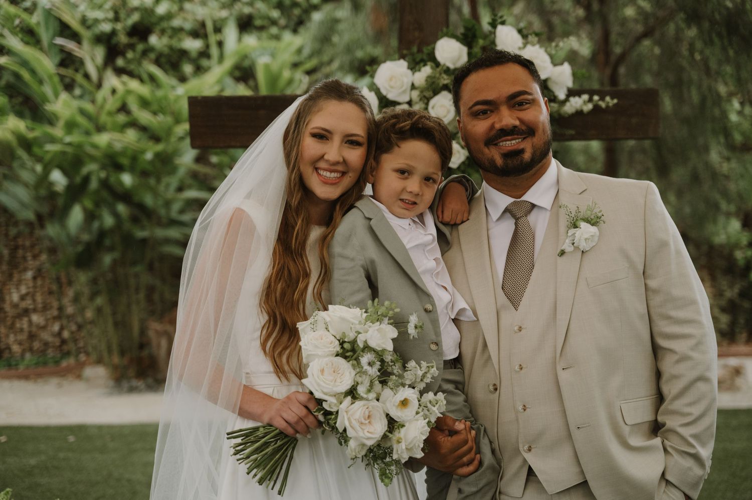 Bride and groom in elegant wedding attire pose with young child in lush garden ceremony setting.