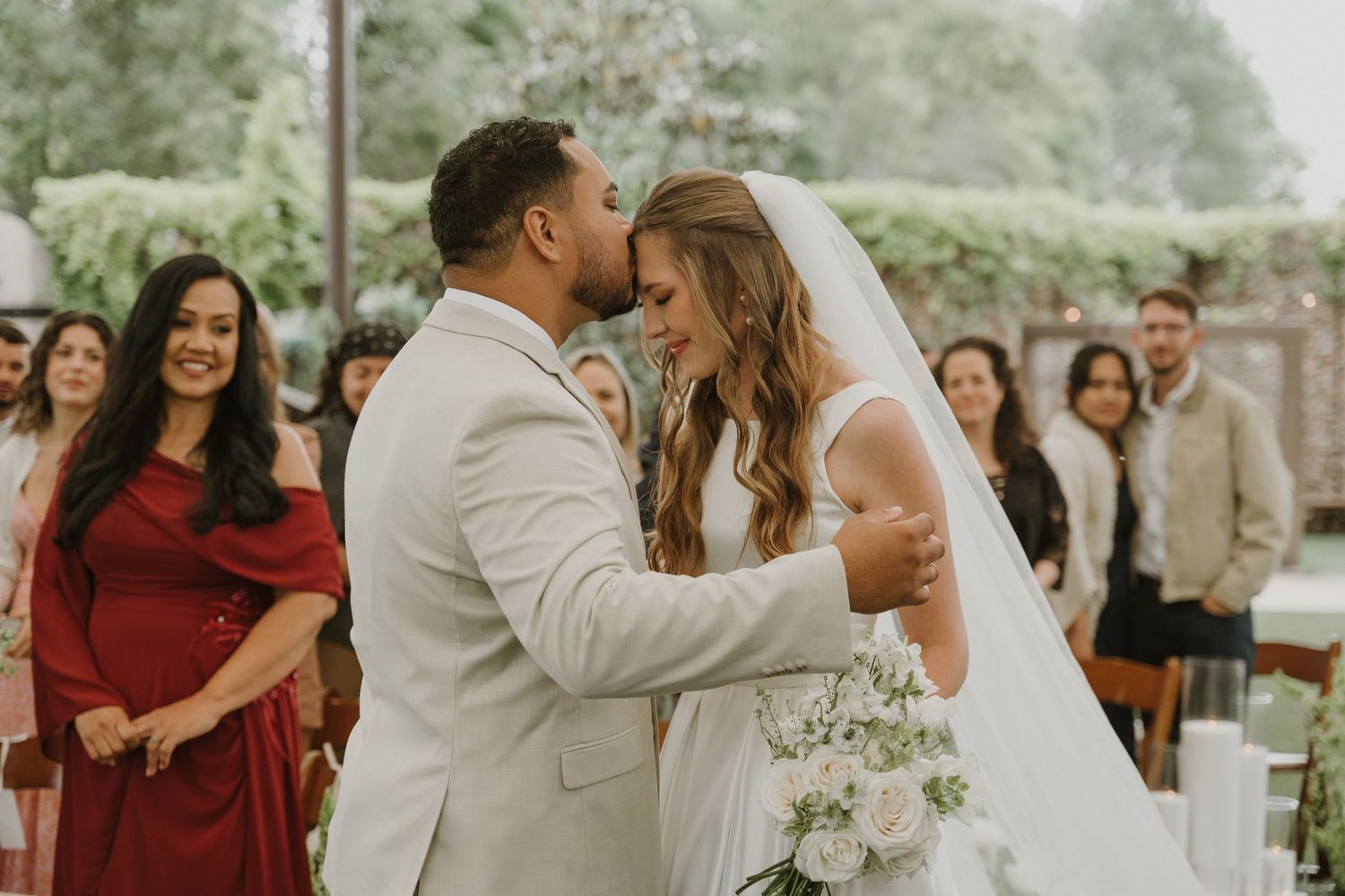 Bride and groom share first kiss at outdoor wedding ceremony with guests watching in background.