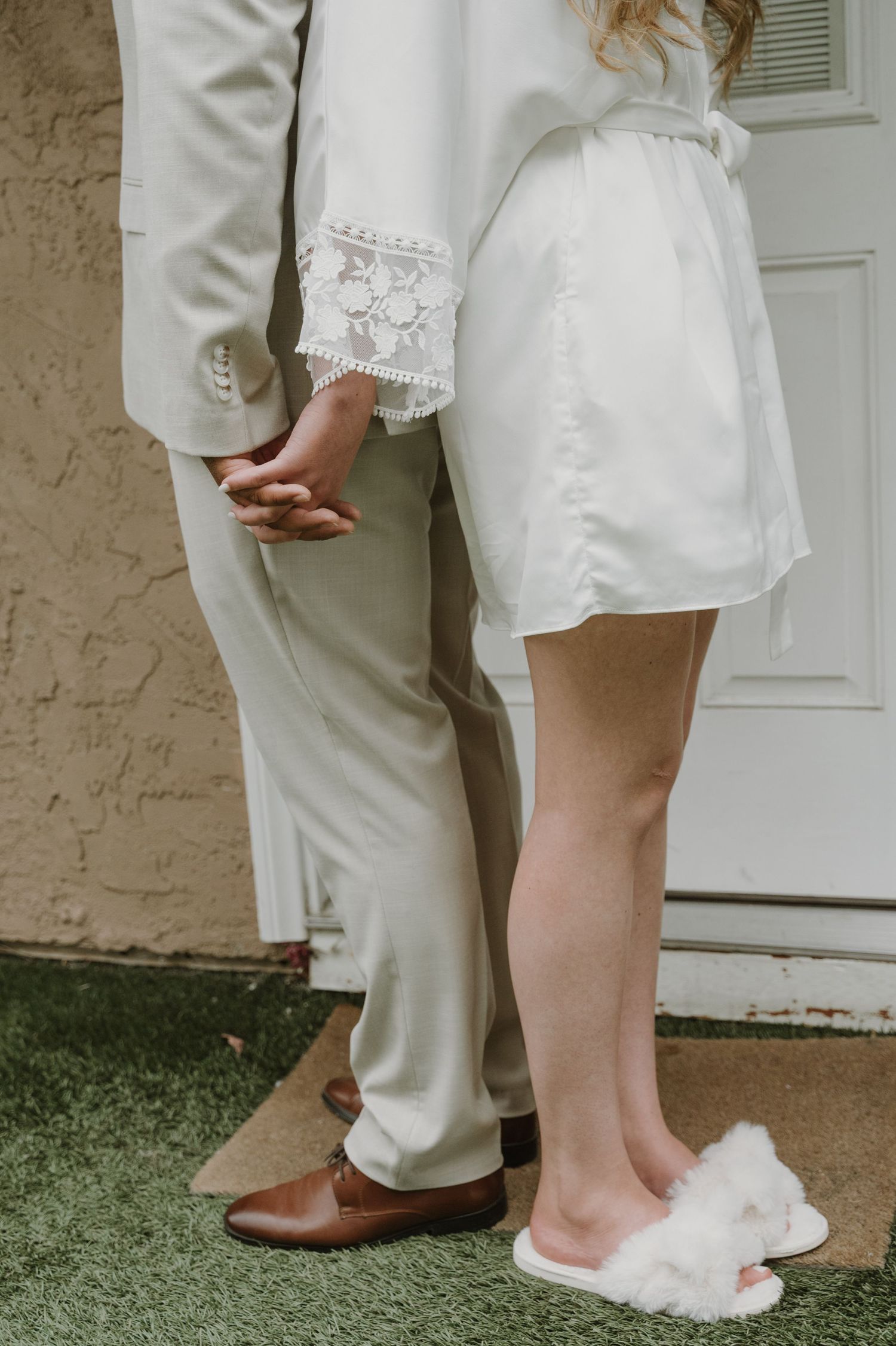 Close-up of bride in white lace robe and fluffy slippers holding hands with groom in beige suit and brown dress shoes.