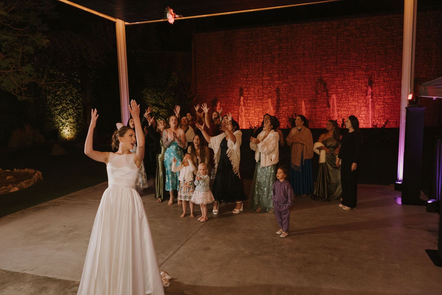 Bride tosses bouquet to excited wedding guests gathered outdoors at night during reception celebration.