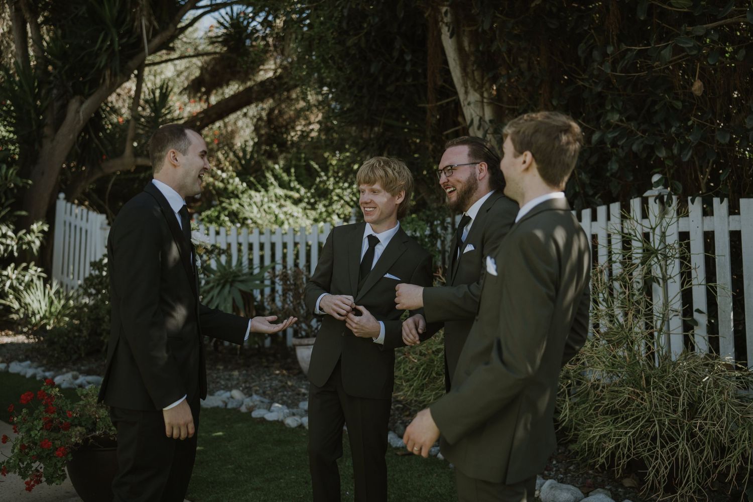 Four groomsmen in dark suits conversing outdoors near a white picket fence and lush garden at a wedding.