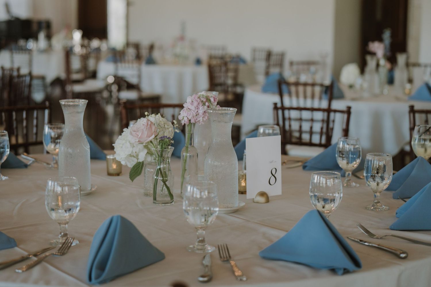 Elegant wedding reception table with blue napkins, floral centerpiece, and glassware on a neutral linen tablecloth.