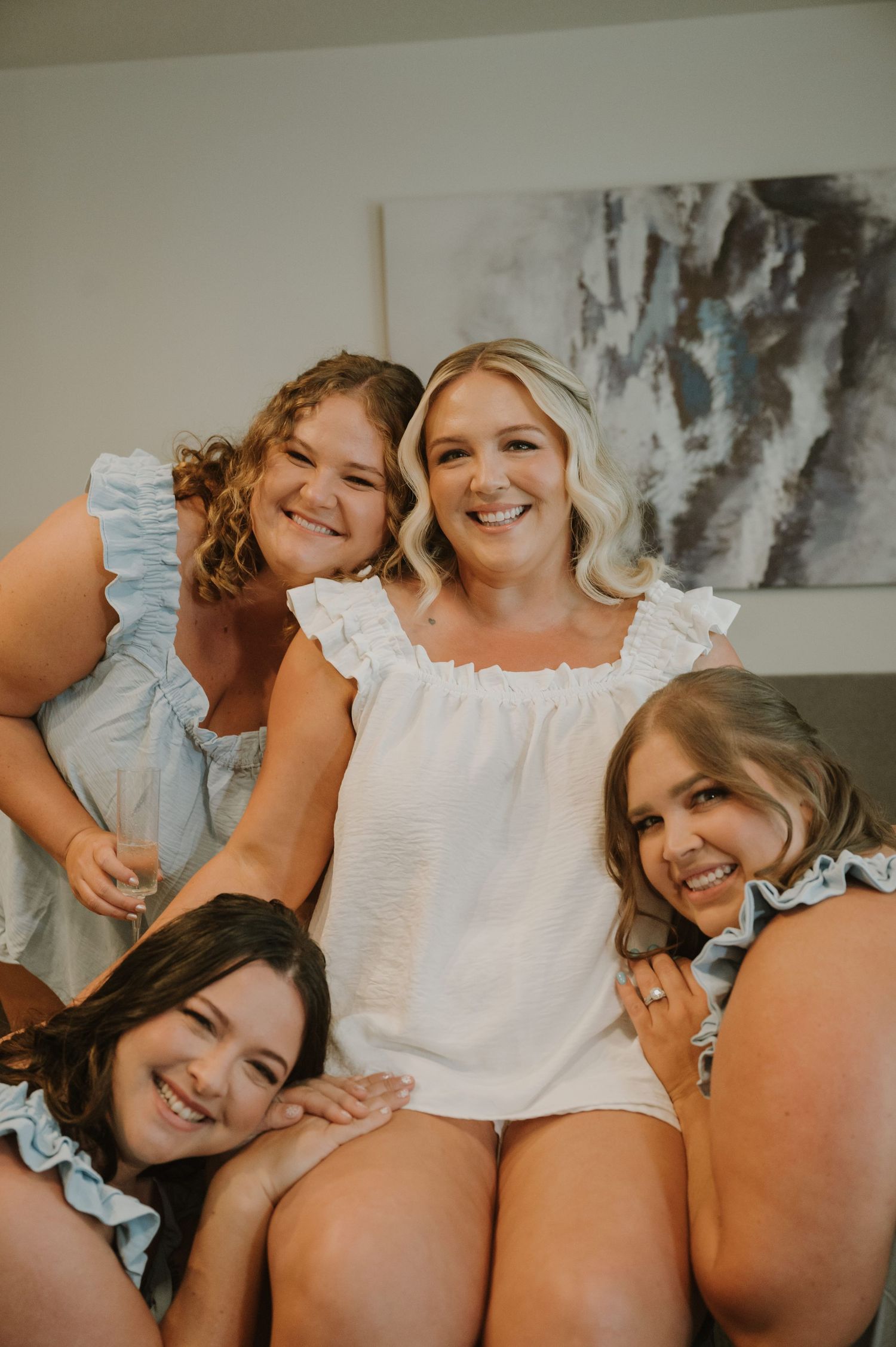 Bride in white top smiling with three bridesmaids in blue ruffled tops during wedding morning preparations.