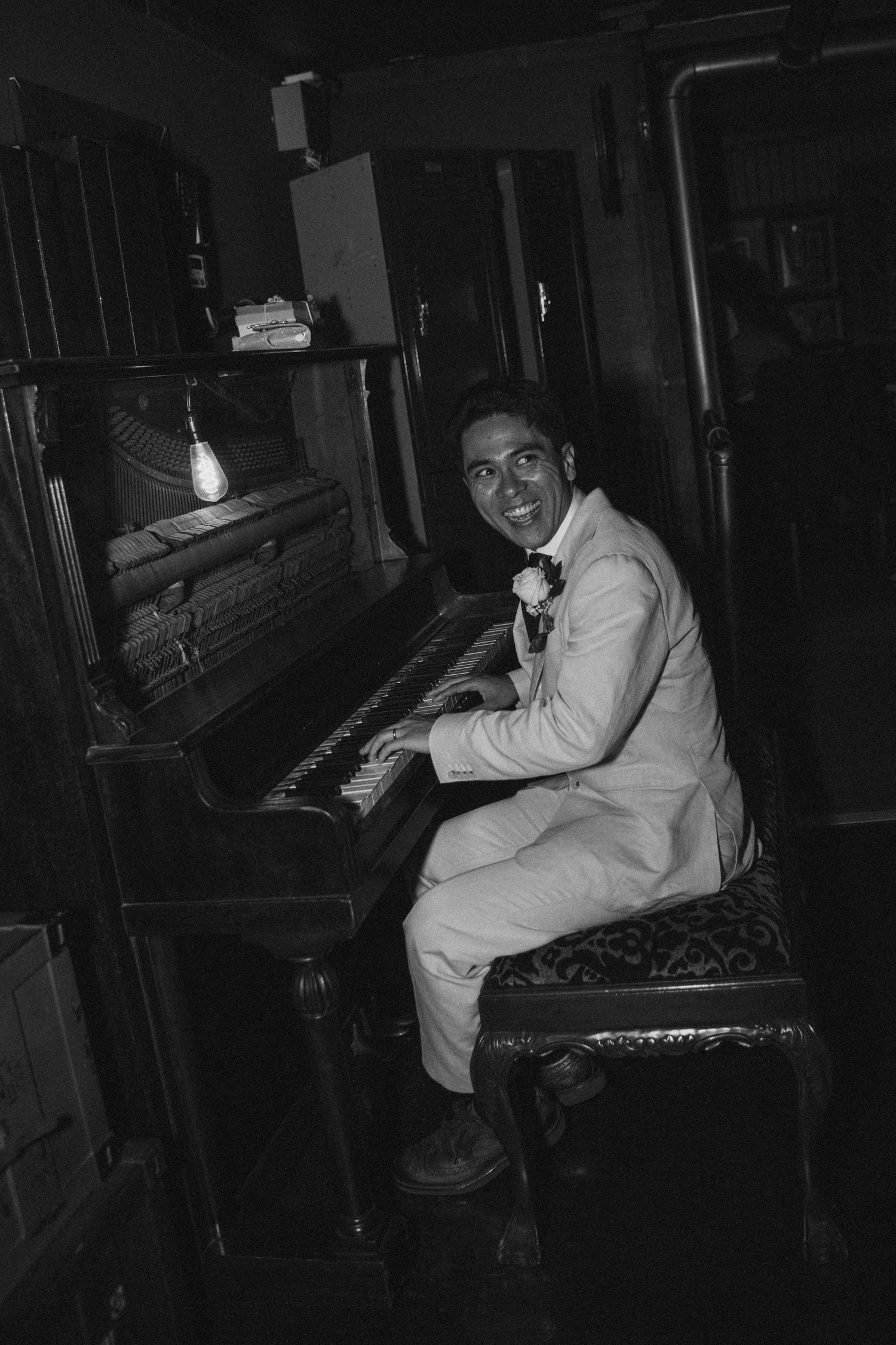 Smiling groom in white suit playing upright piano in dimly lit vintage room, black and white photo.