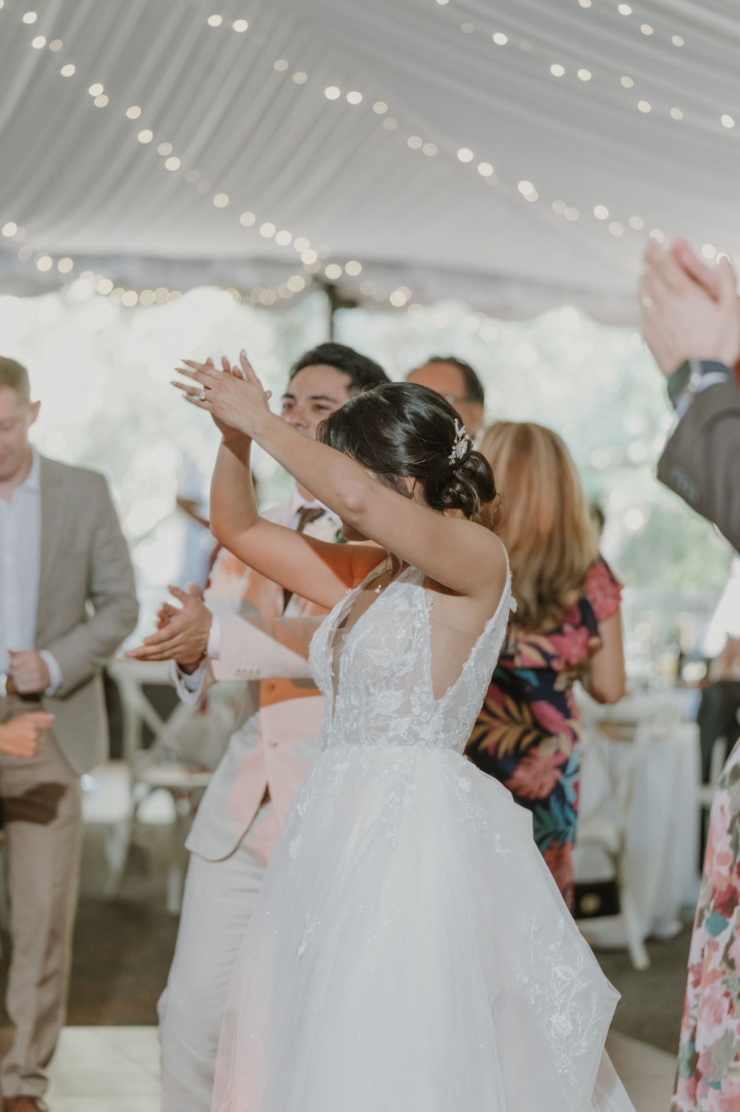 Bride and groom dance joyfully with raised hands among guests at tented wedding reception.