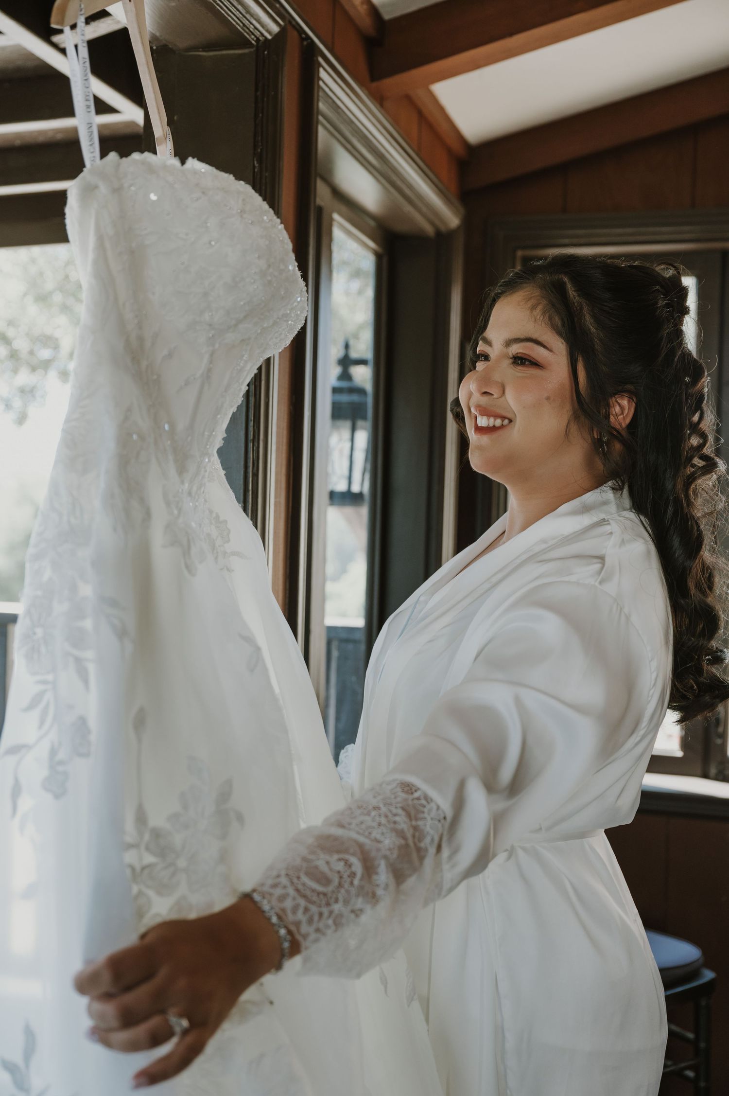 Smiling bride in white lace robe admiring her strapless wedding gown hanging by a window before her ceremony.