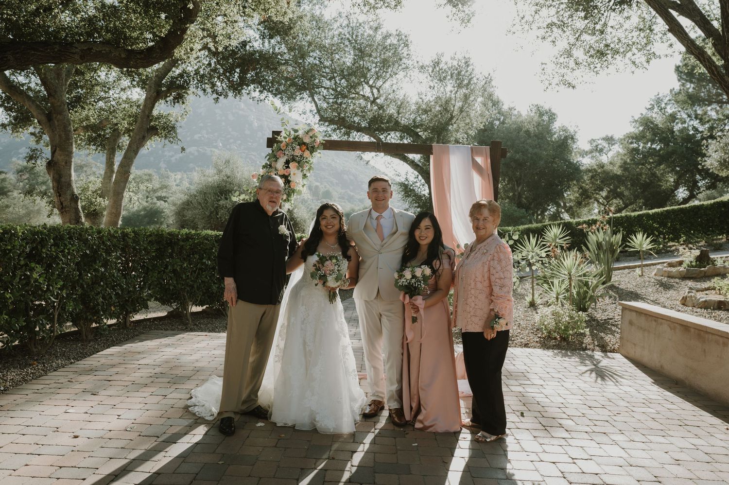 Wedding couple poses with family members under a floral arch at an outdoor garden ceremony venue.