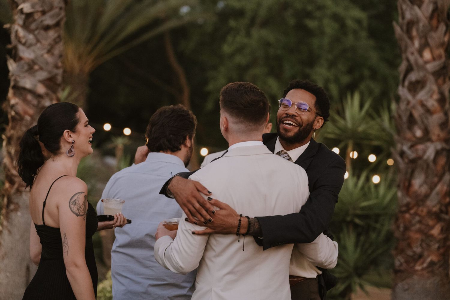 Guests hugging and laughing outdoors at a wedding reception surrounded by palm trees and string lights.