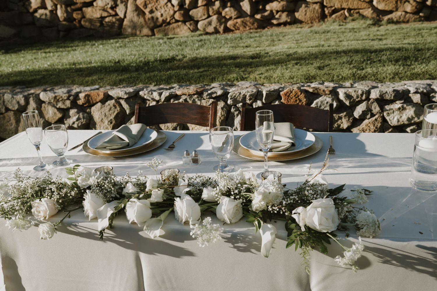 An elegant white table setting with floral centerpieces and place settings against a stone wall.