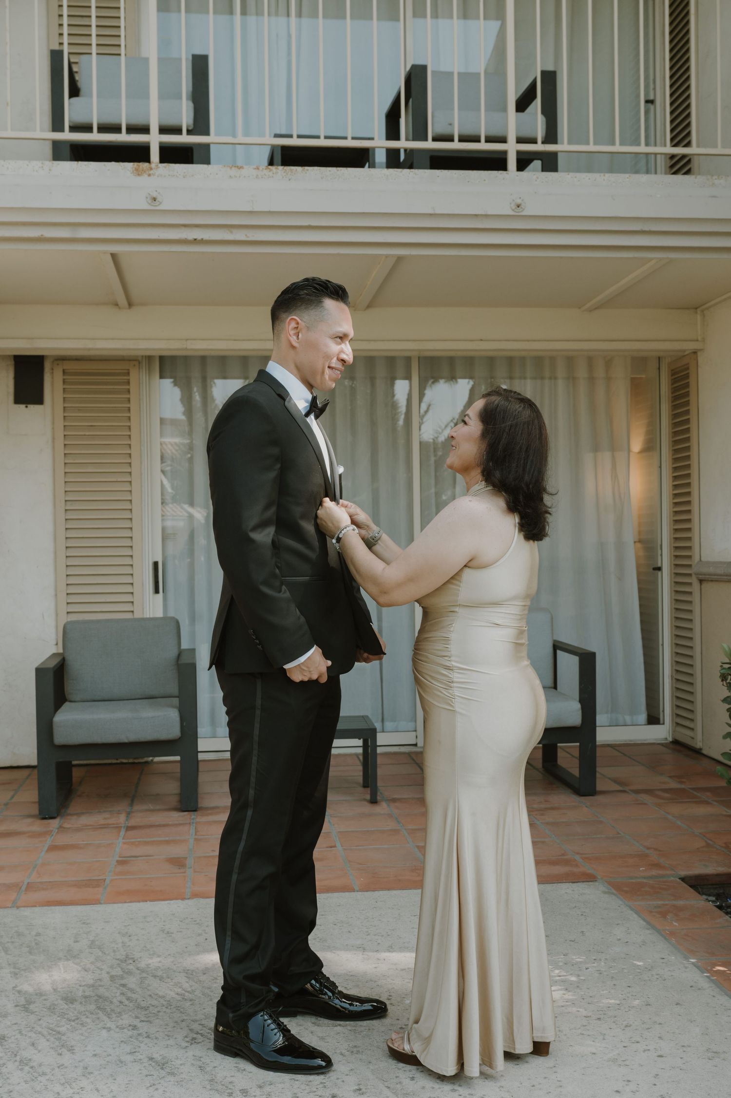 A couple shares a candid moment while adjusting formal wedding attire on a patio.