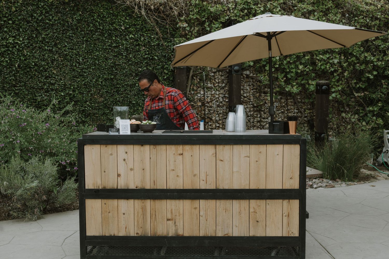 Man working at an outdoor wooden bar cart with umbrella, surrounded by lush green ivy wall backdrop.