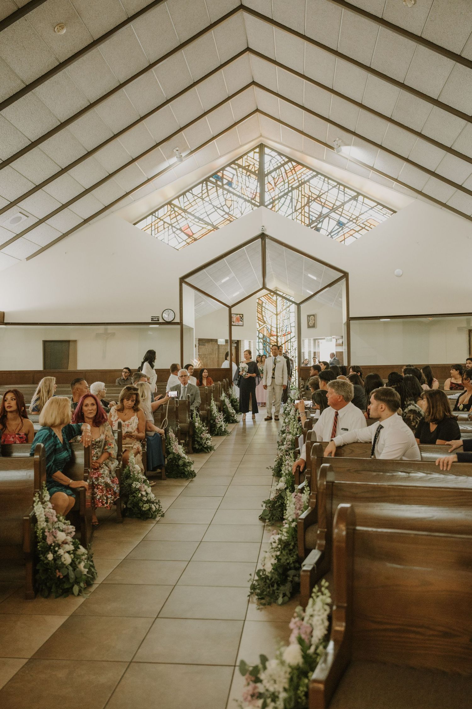 Floral-decorated church aisle with wedding guests seated in wooden pews beneath stained glass skylights.
