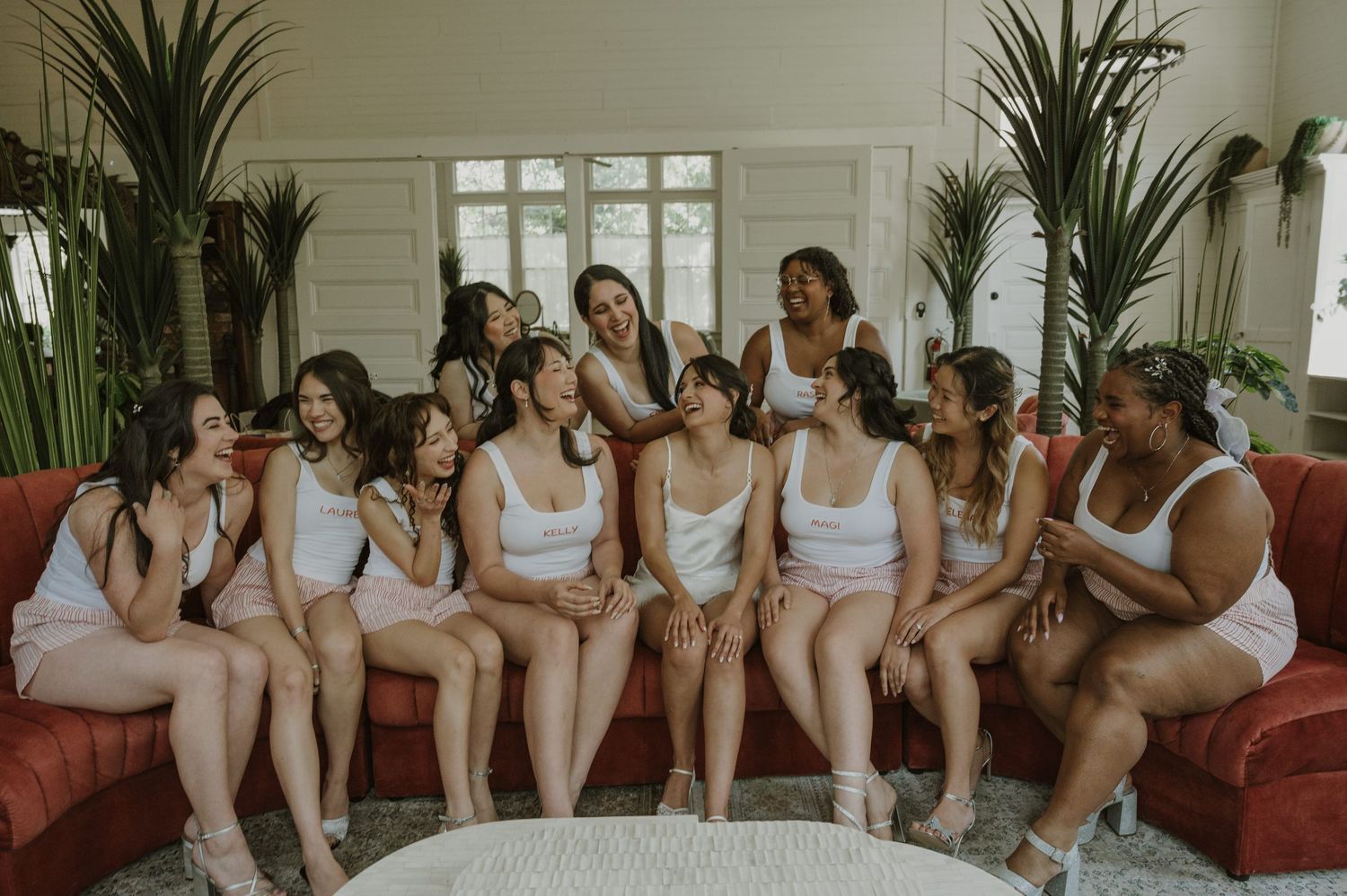 Group of bridesmaids in white loungewear laughing and sitting together on a red sofa before the wedding.