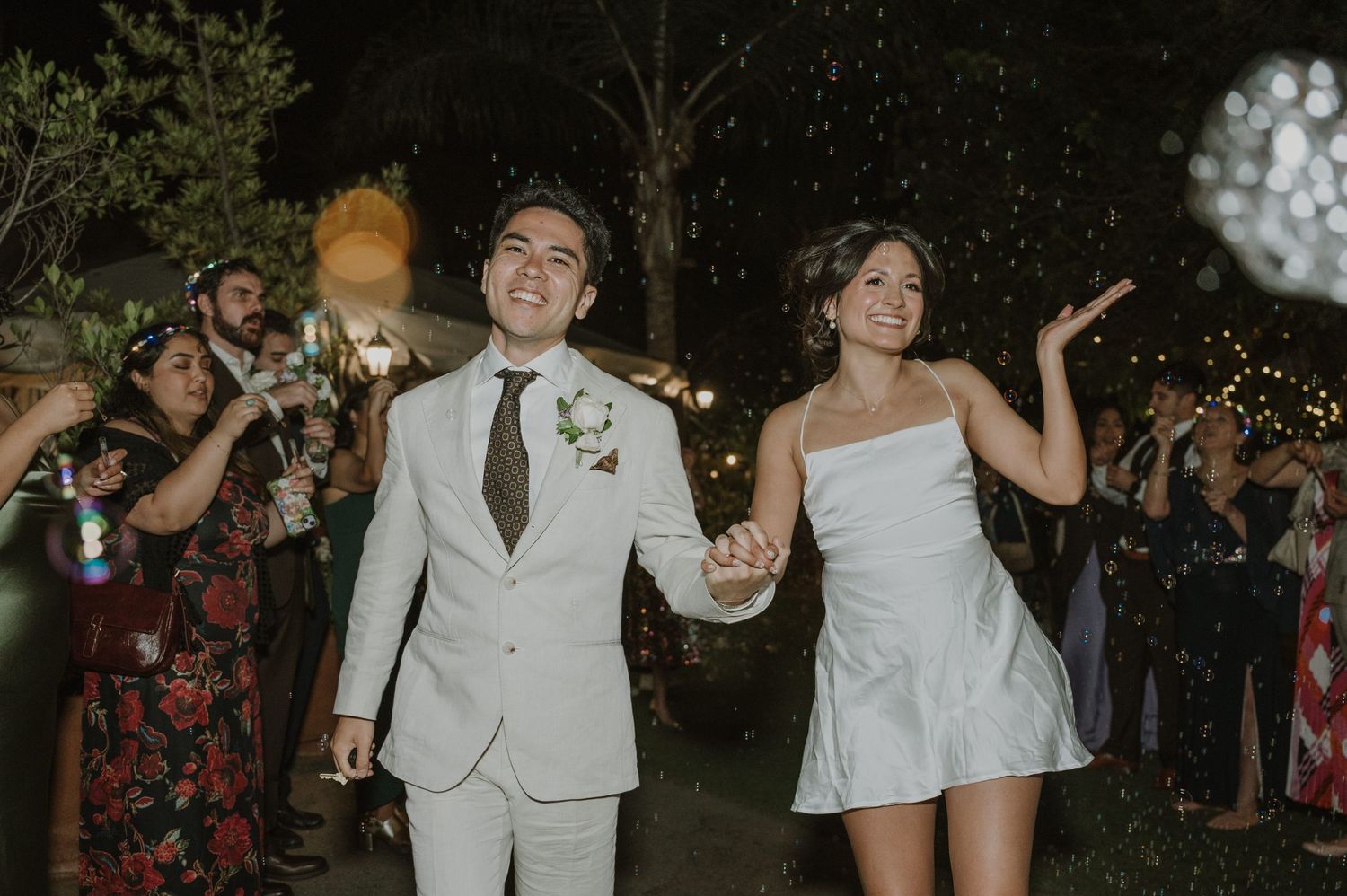 Newlyweds walk hand-in-hand through sparkler send-off at outdoor evening wedding celebration.