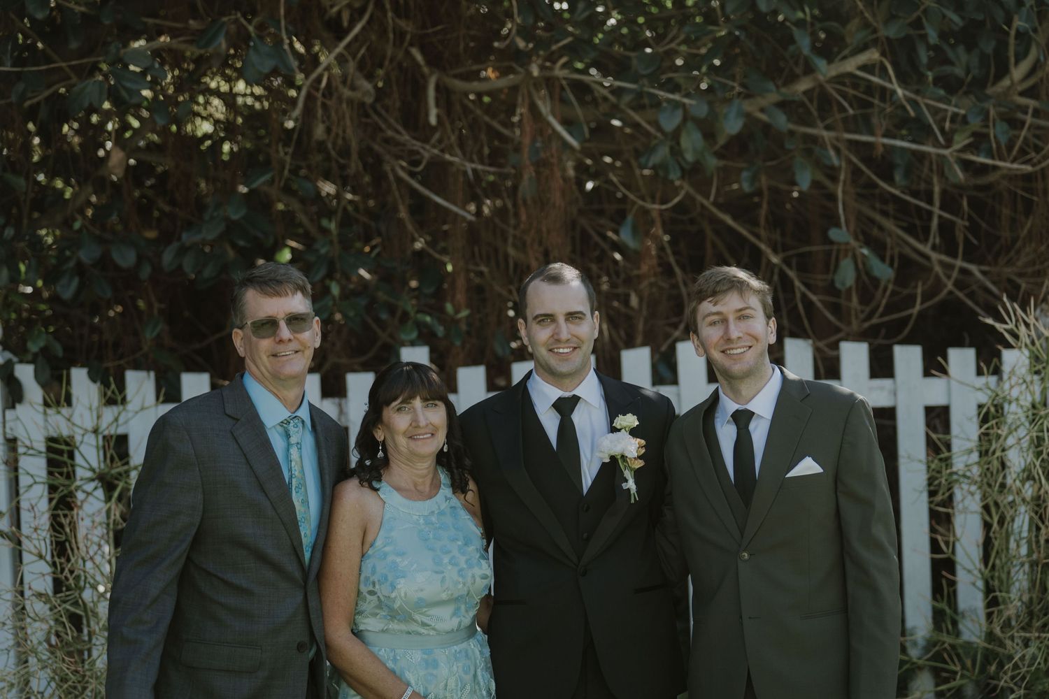 Family of four posing together outdoors at a wedding, dressed in formal attire near a white picket fence.