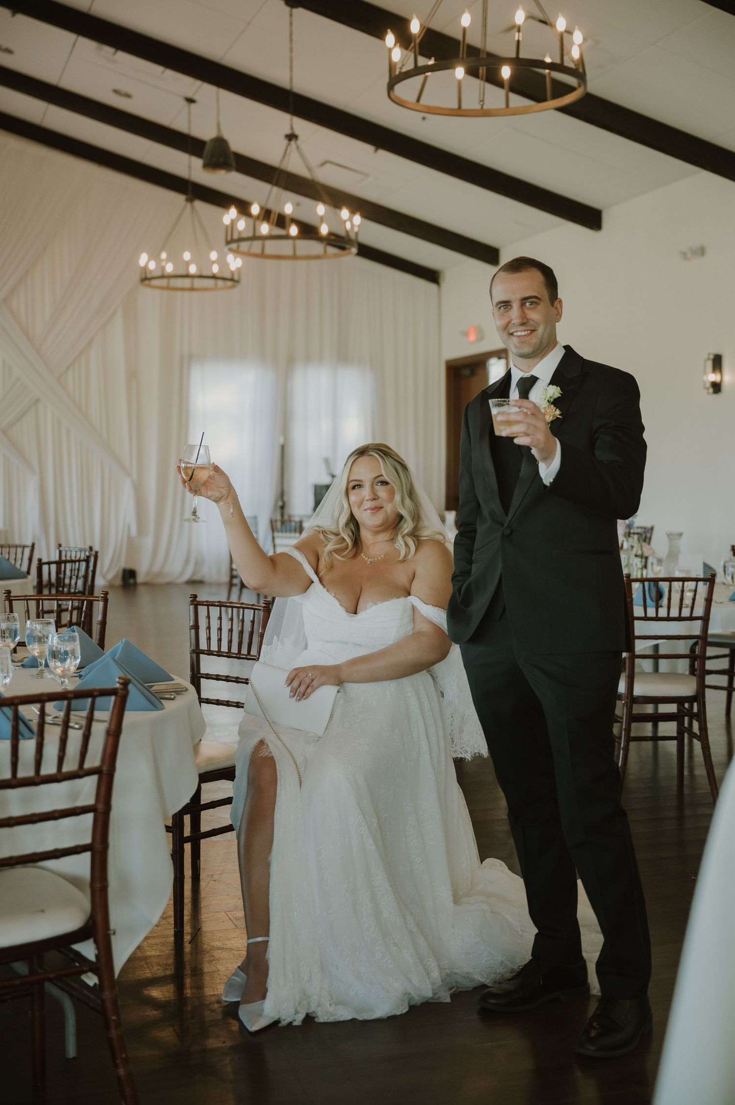 Bride and groom raising champagne glasses in elegant wedding reception hall with chandelier lighting.