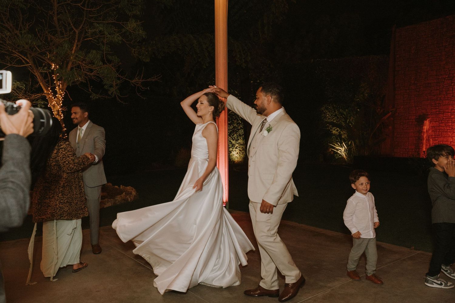 Groom in beige suit twirling bride in white satin gown during outdoor evening wedding reception dance celebration.