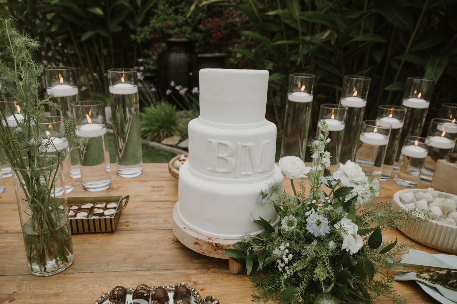 White two-tier wedding cake with monogram initials surrounded by floating candles and floral arrangements on a wooden table.