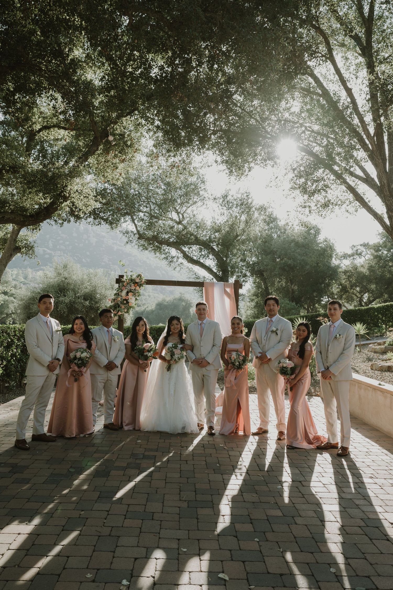 Wedding party poses in pink dresses and white suits under trees at an outdoor ceremony.