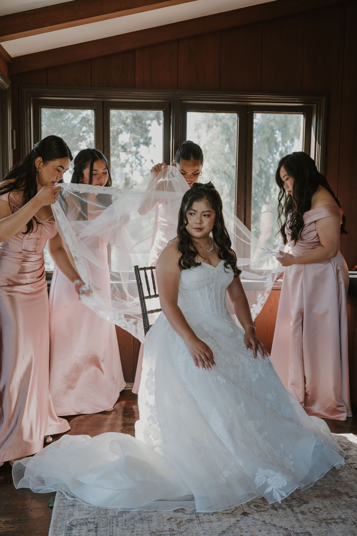 Bridesmaids in pink dresses arrange the veil of a bride in a white lace wedding gown.