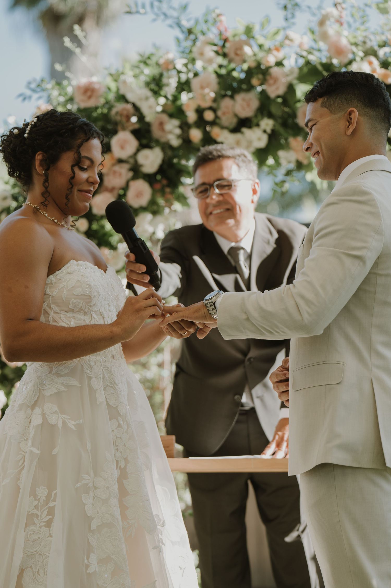 Bride placing wedding ring on groom's finger during outdoor ceremony with floral arch backdrop.