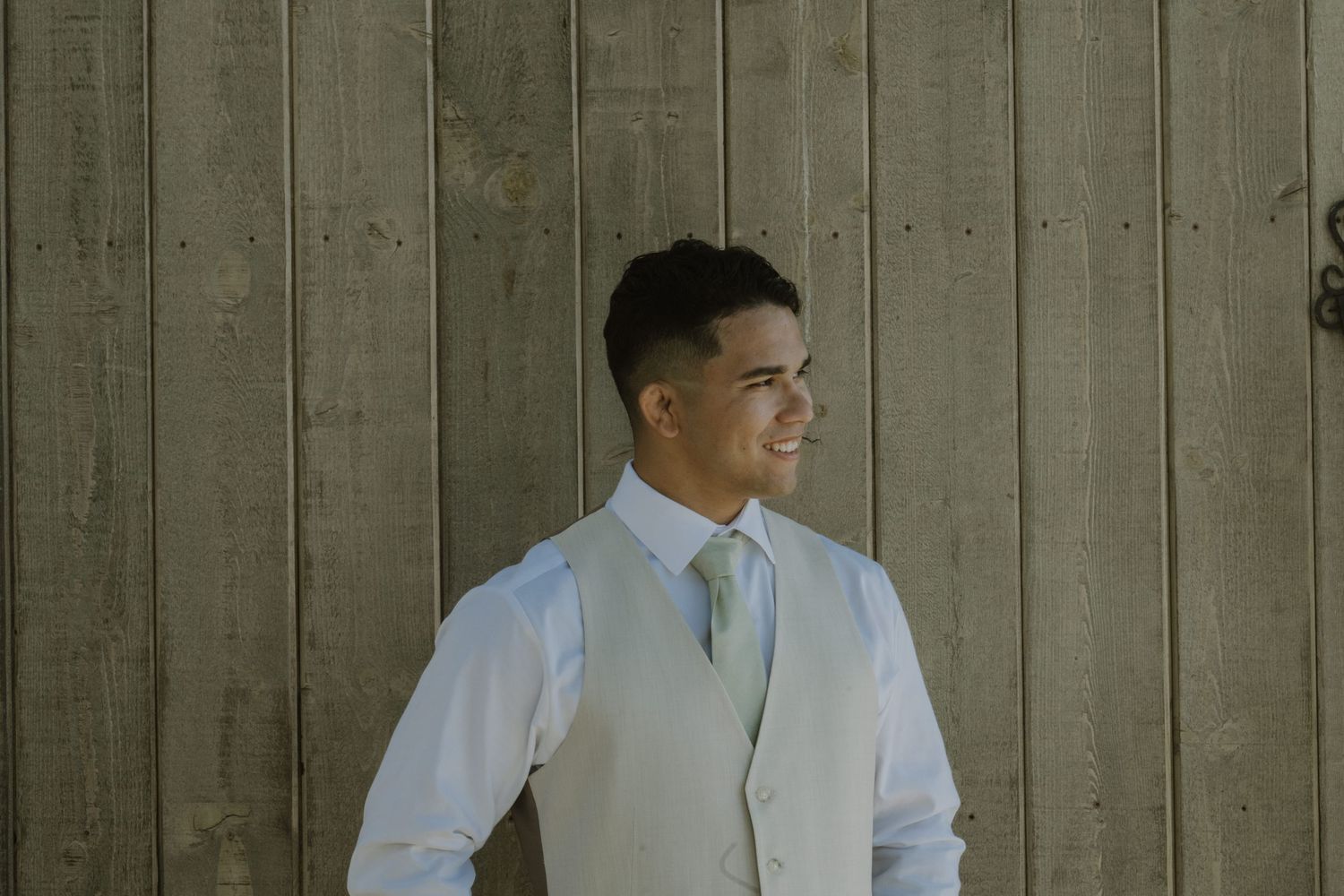 Handsome groom in white vest and tie smiles while standing against a rustic wooden wall.
