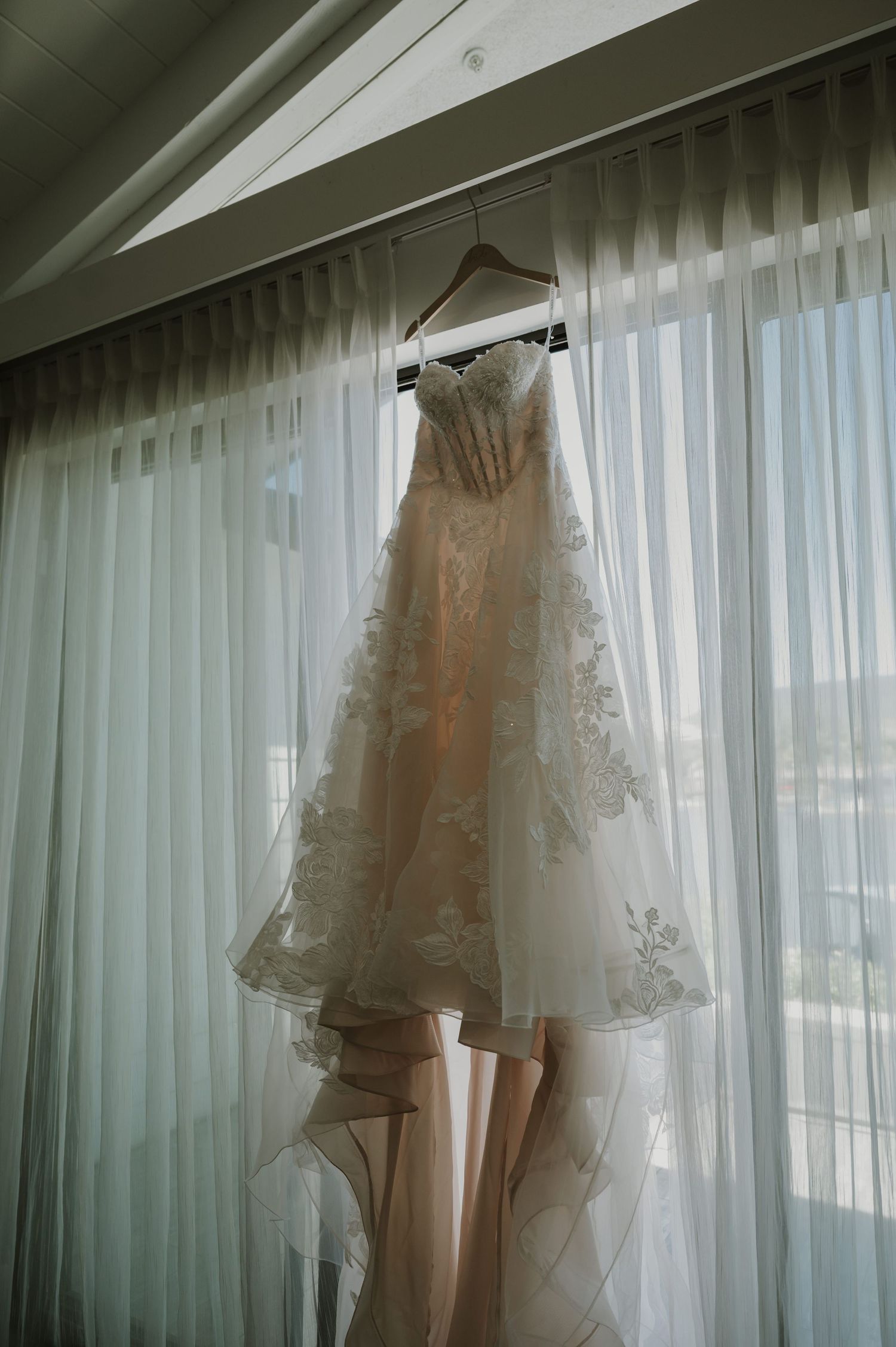 Elegant lace wedding dress hanging on a hanger against sheer white curtains in soft natural light.