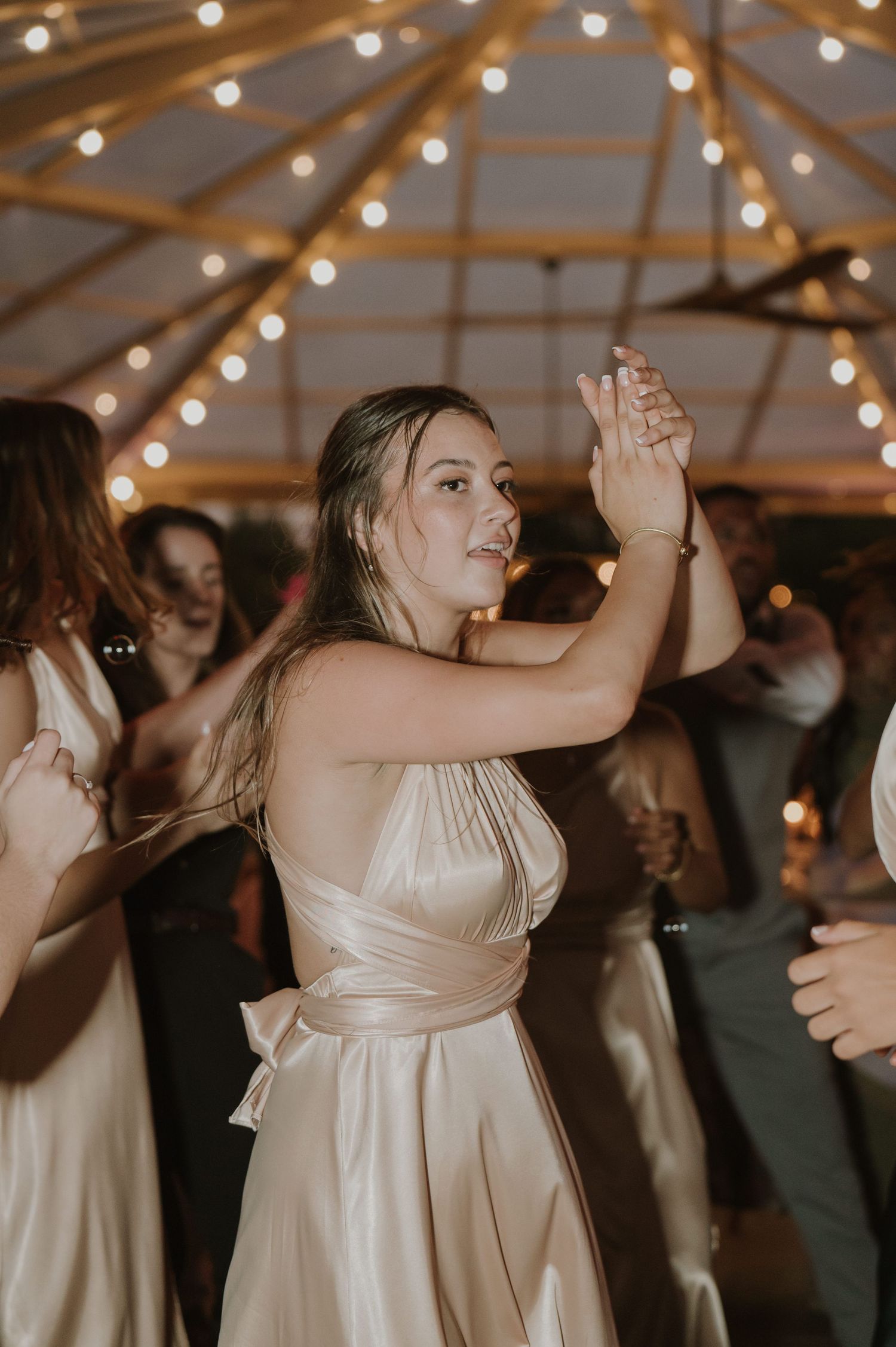 A bridesmaid in a champagne satin dress dances joyfully under string lights at a wedding reception.