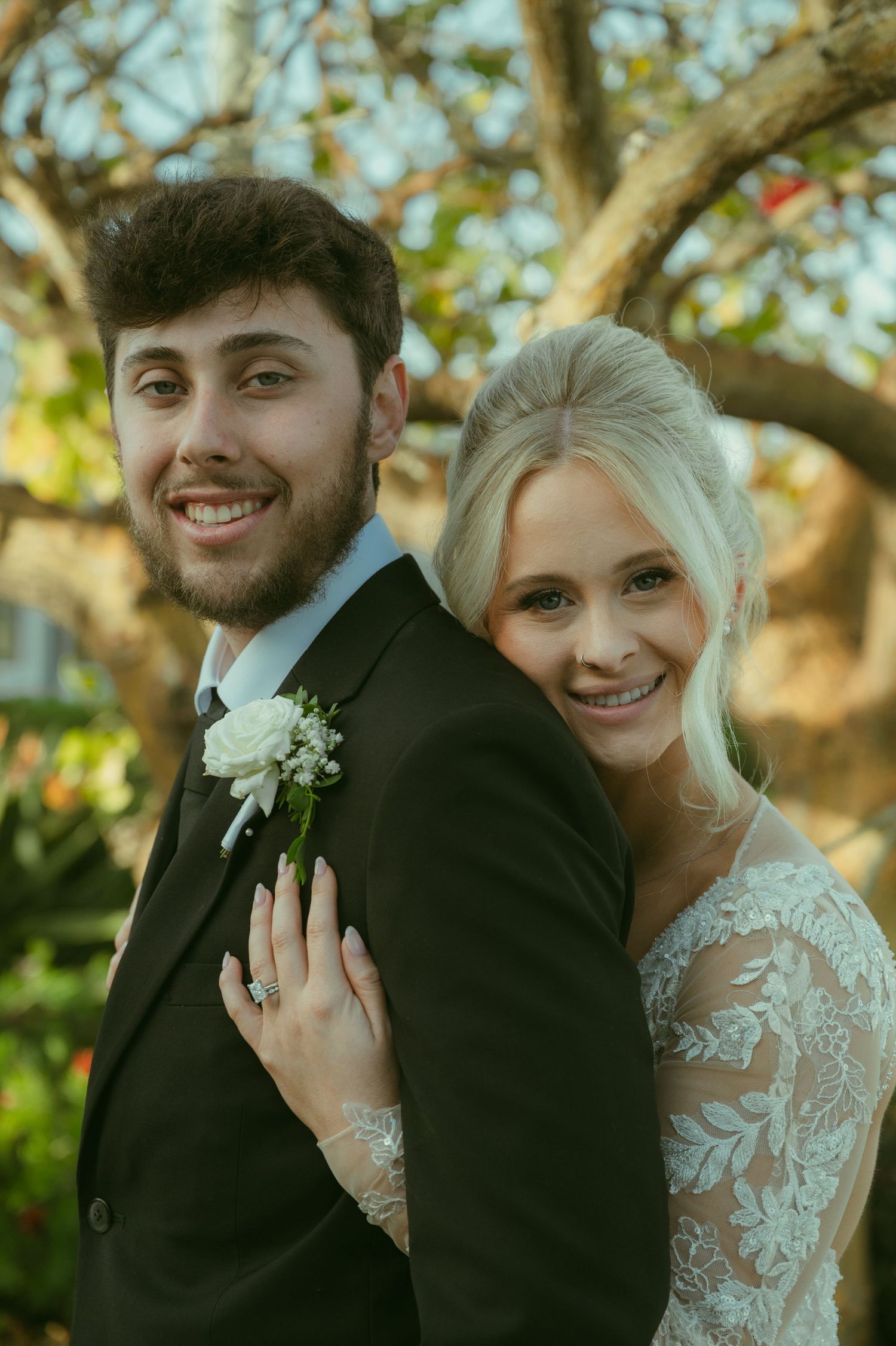 Smiling bride and groom pose together outdoors, she in lace wedding gown, he in dark suit with white boutonniere.