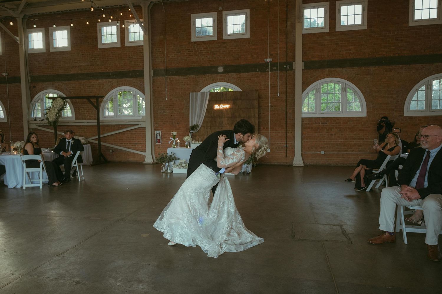 Bride and groom share romantic first dance dip in elegant brick wedding venue with white chairs and guests.