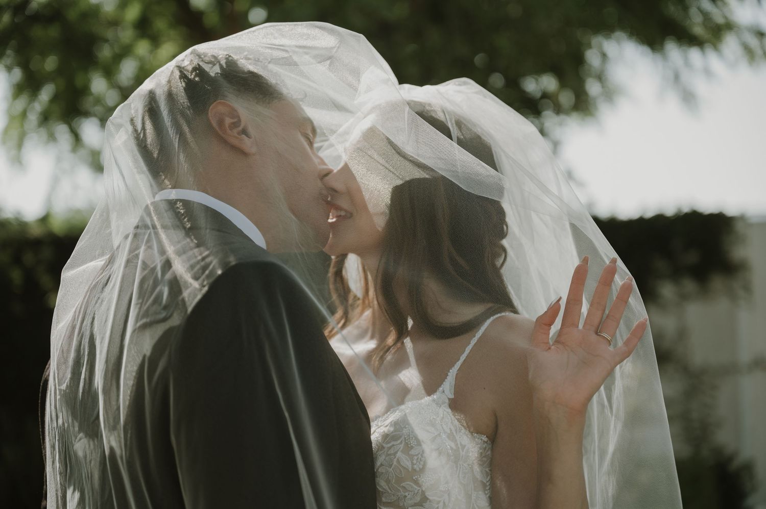 Bride and groom sharing a romantic kiss under a flowing white wedding veil outdoors.